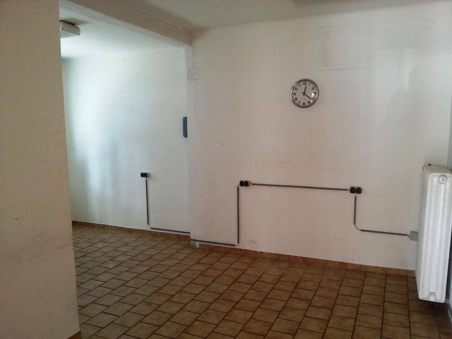 Leerer Raum mit Fliesenboden, Heizkörper und Wanduhr, mit sichtbaren Installationen.