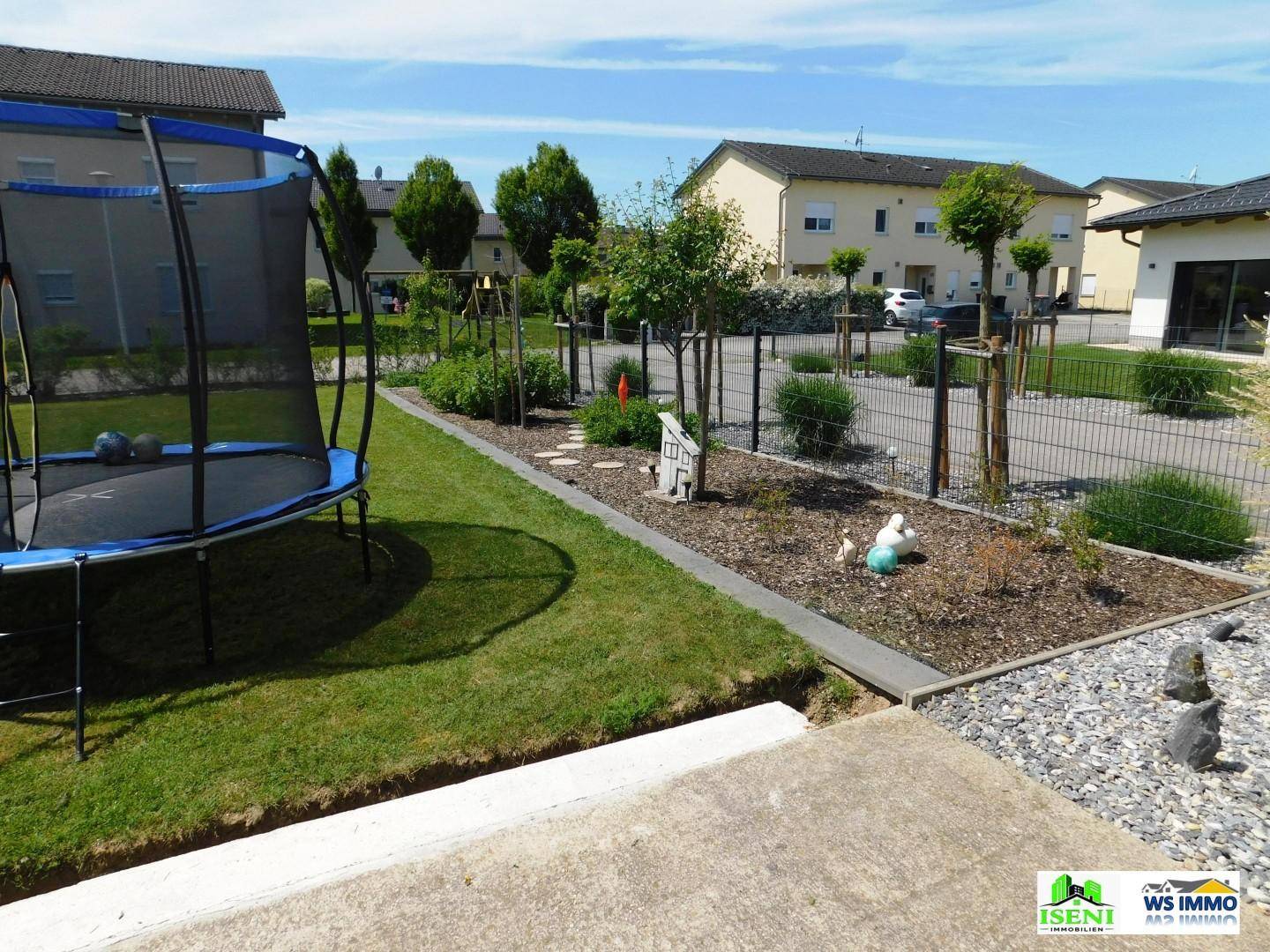 Gestalteter Gartenbereich mit Trampolin, Zierbeeten und einem kleinen Weg aus Trittsteinen.