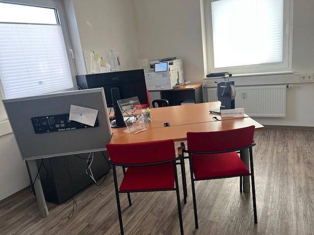 Helles Büro mit Schreibtisch, Computermonitor und zwei roten Stühlen vor einem Fenster.