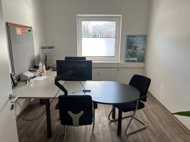 Helles Einzelbüro mit Schreibtisch, Computer und einem runden Besprechungstisch am Fenster.