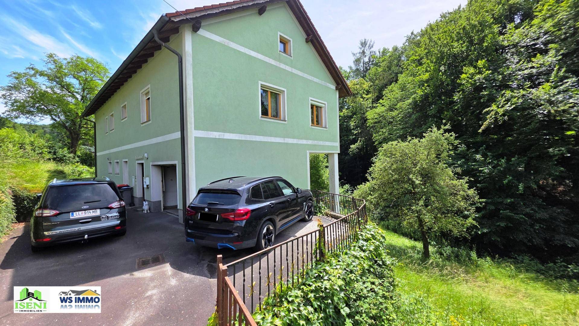 Grünes Einfamilienhaus mit Satteldach, umgeben von Bäumen und zwei Autos auf der Auffahrt.