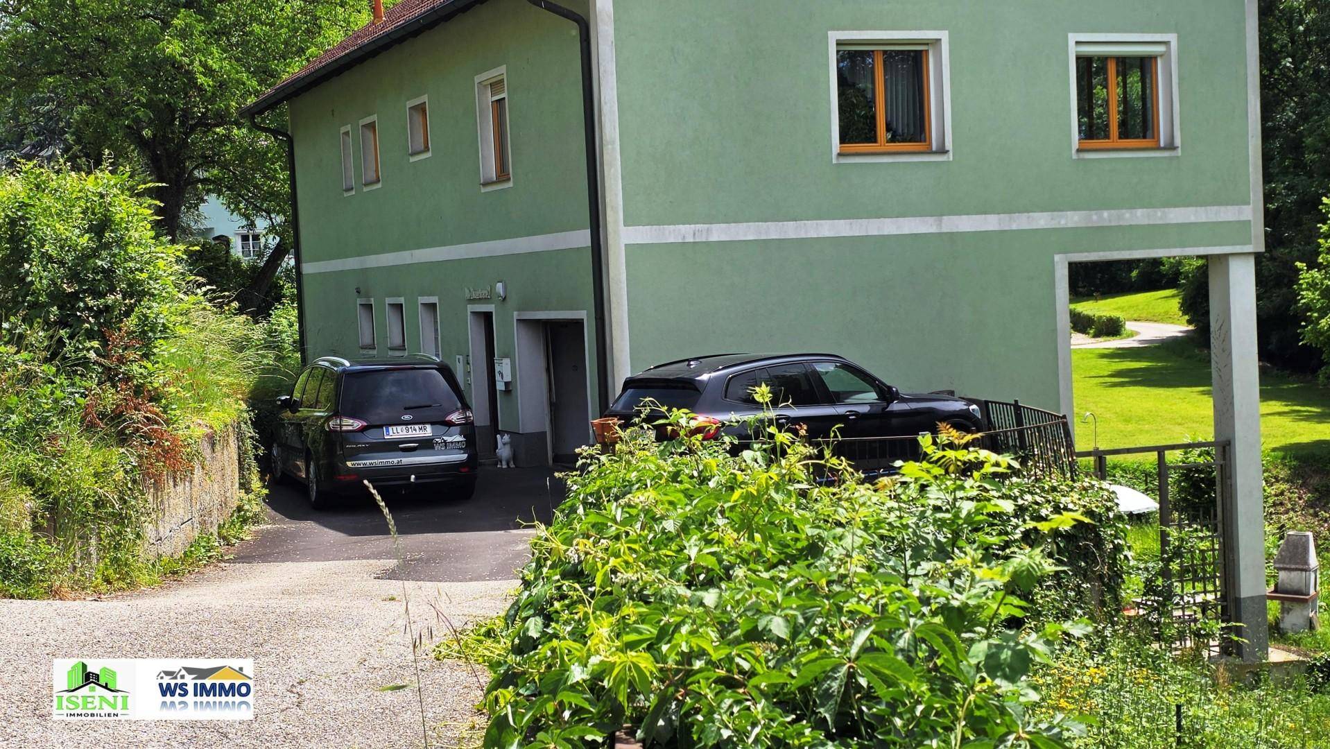Grünes Einfamilienhaus mit Satteldach, umgeben von Bäumen und zwei Autos auf der Auffahrt.