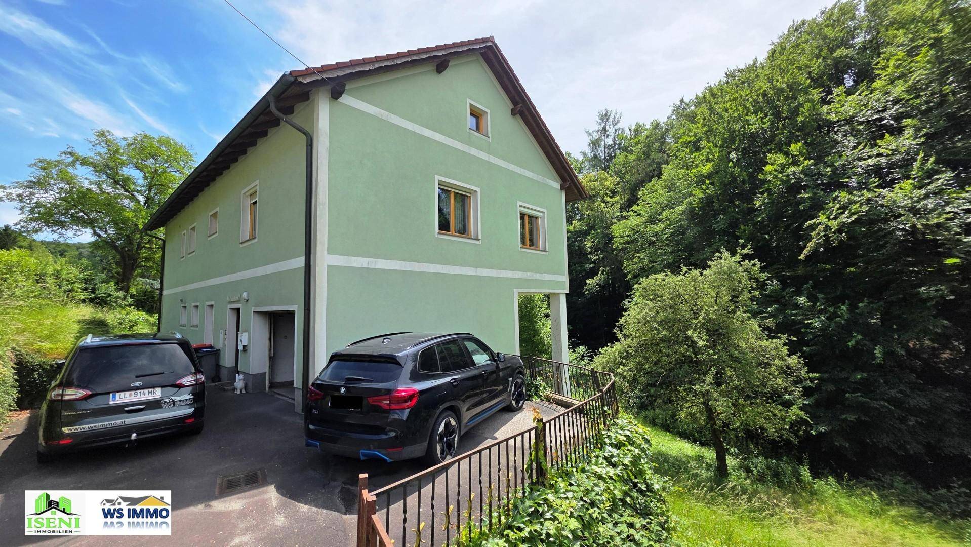 Grünes Einfamilienhaus mit Satteldach, umgeben von Bäumen und zwei Autos auf der Auffahrt.