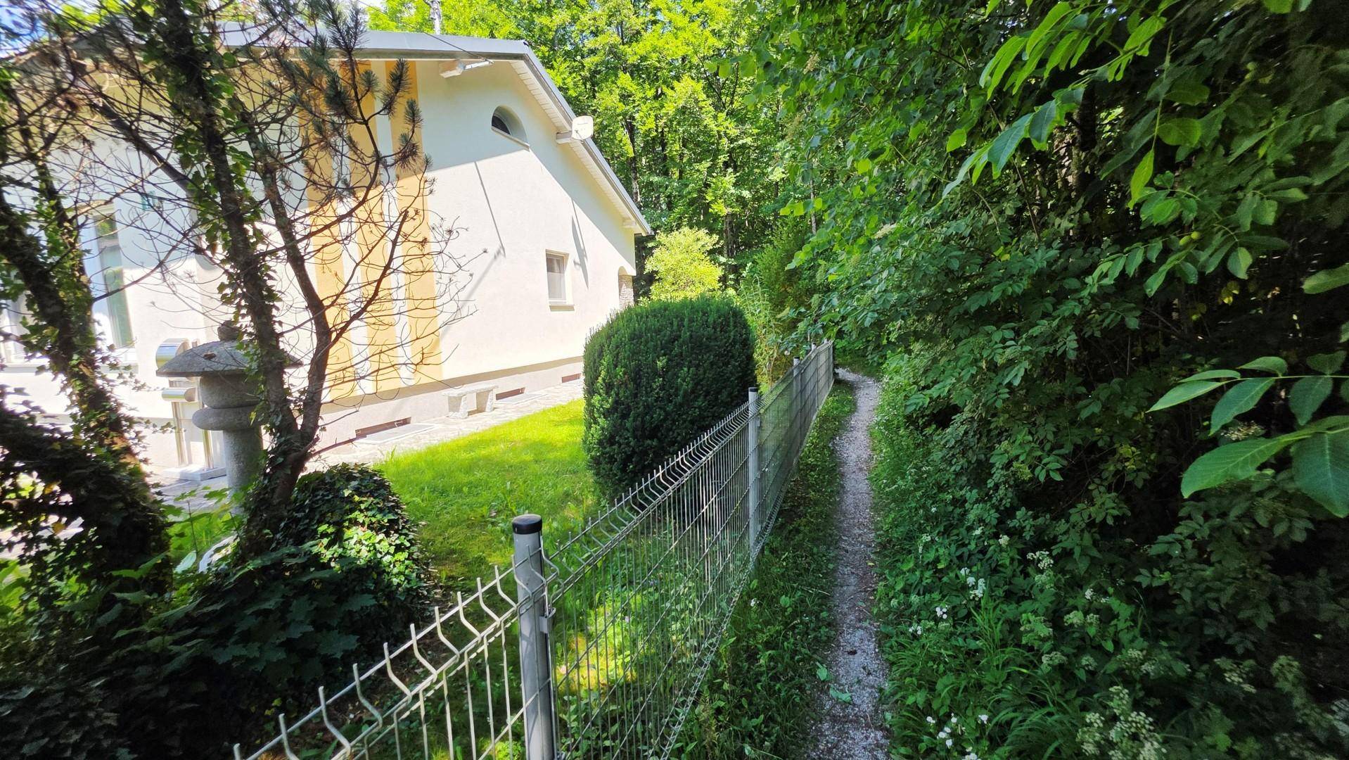 Seitenansicht des Hauses mit einem schmalen Weg und Zaun, der an einen Wald grenzt.