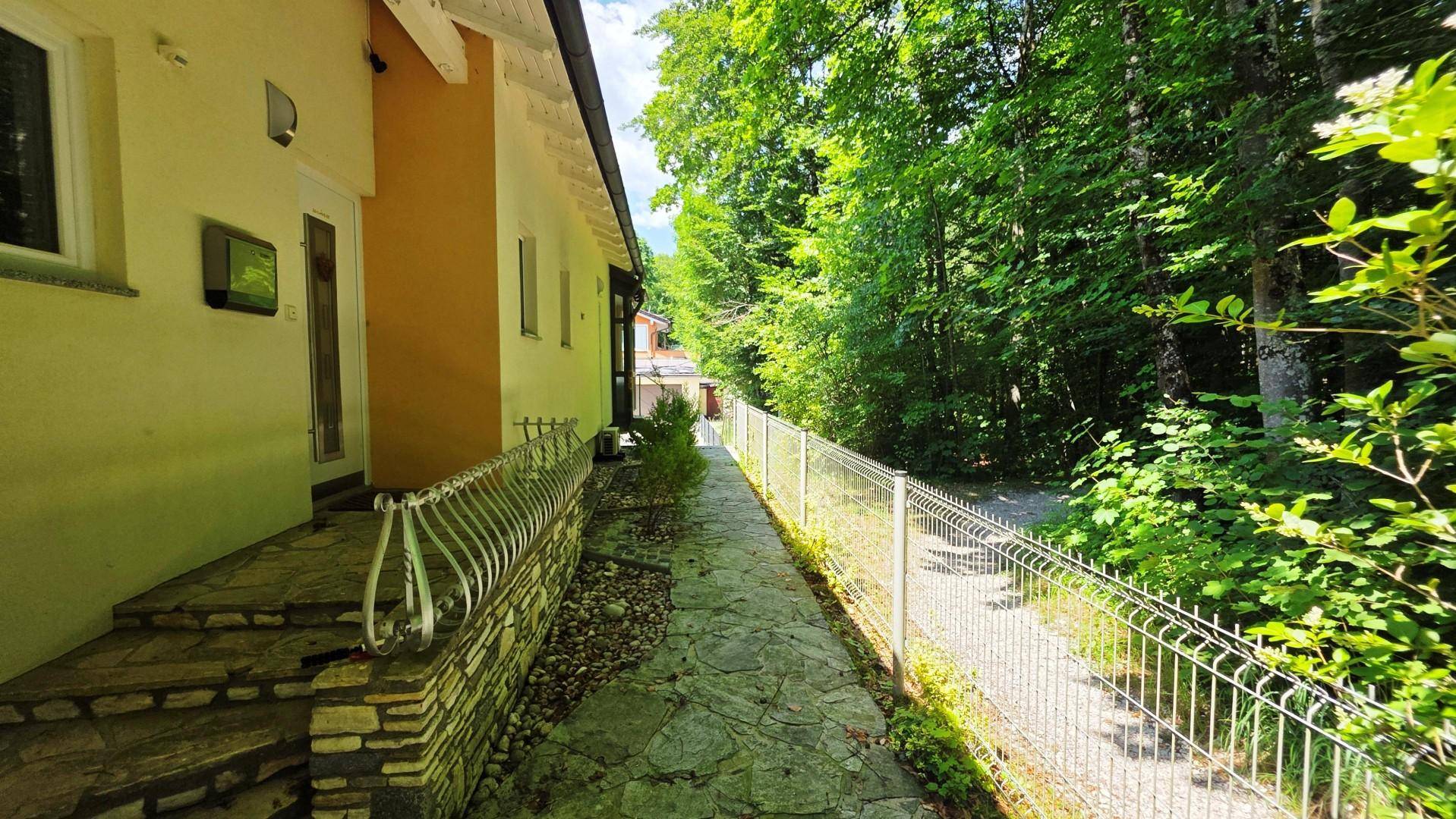 Schmaler Steinweg entlang der Hauswand und eines Zauns, der zum Wald führt.