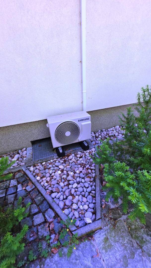 Außenansicht einer Wärmepumpe an der Hauswand, umgeben von Kies und Pflanzen.