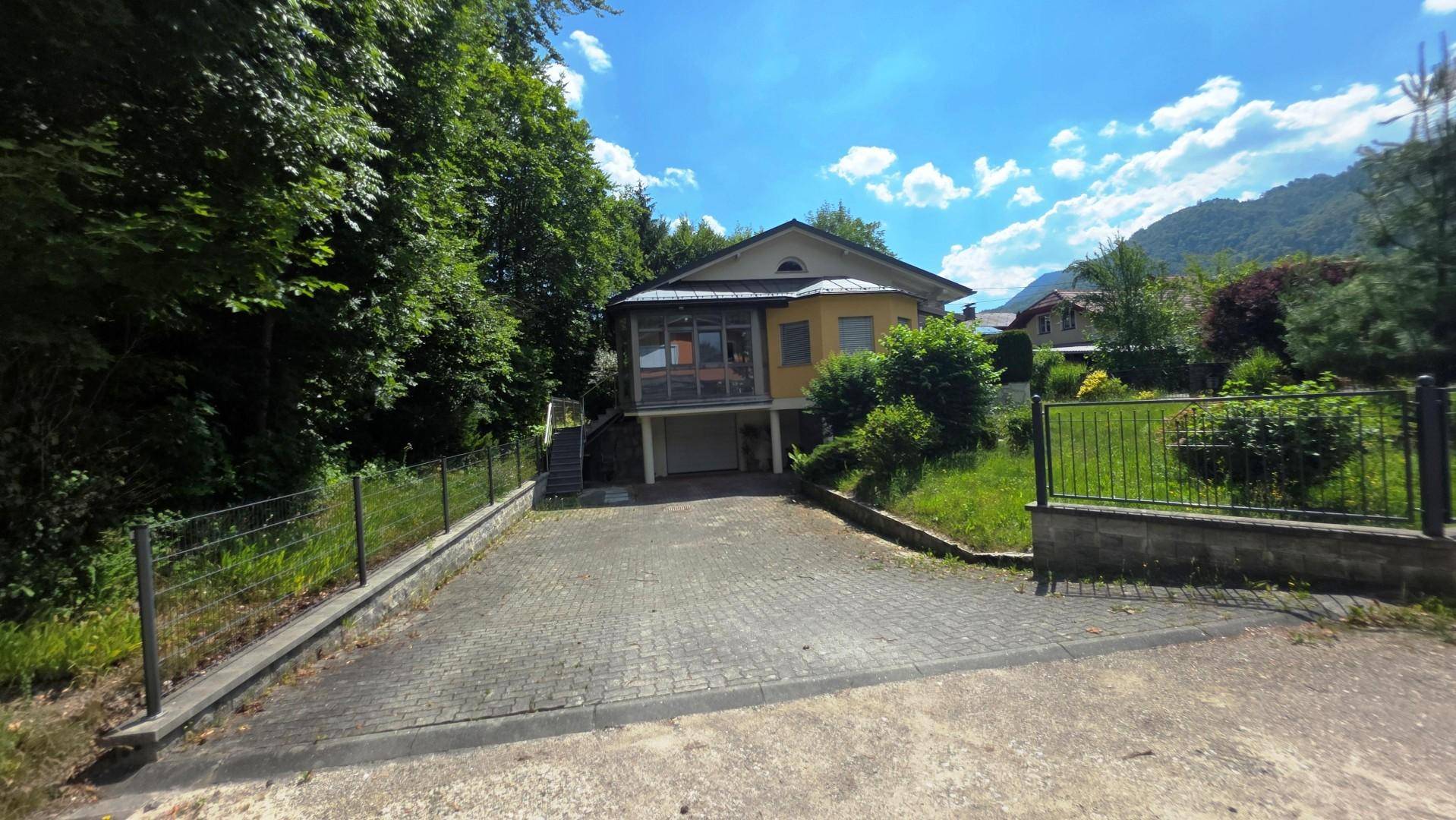 Ansicht des gelben Hauses mit Garage, gepflasterter Auffahrt und umgebender Natur.