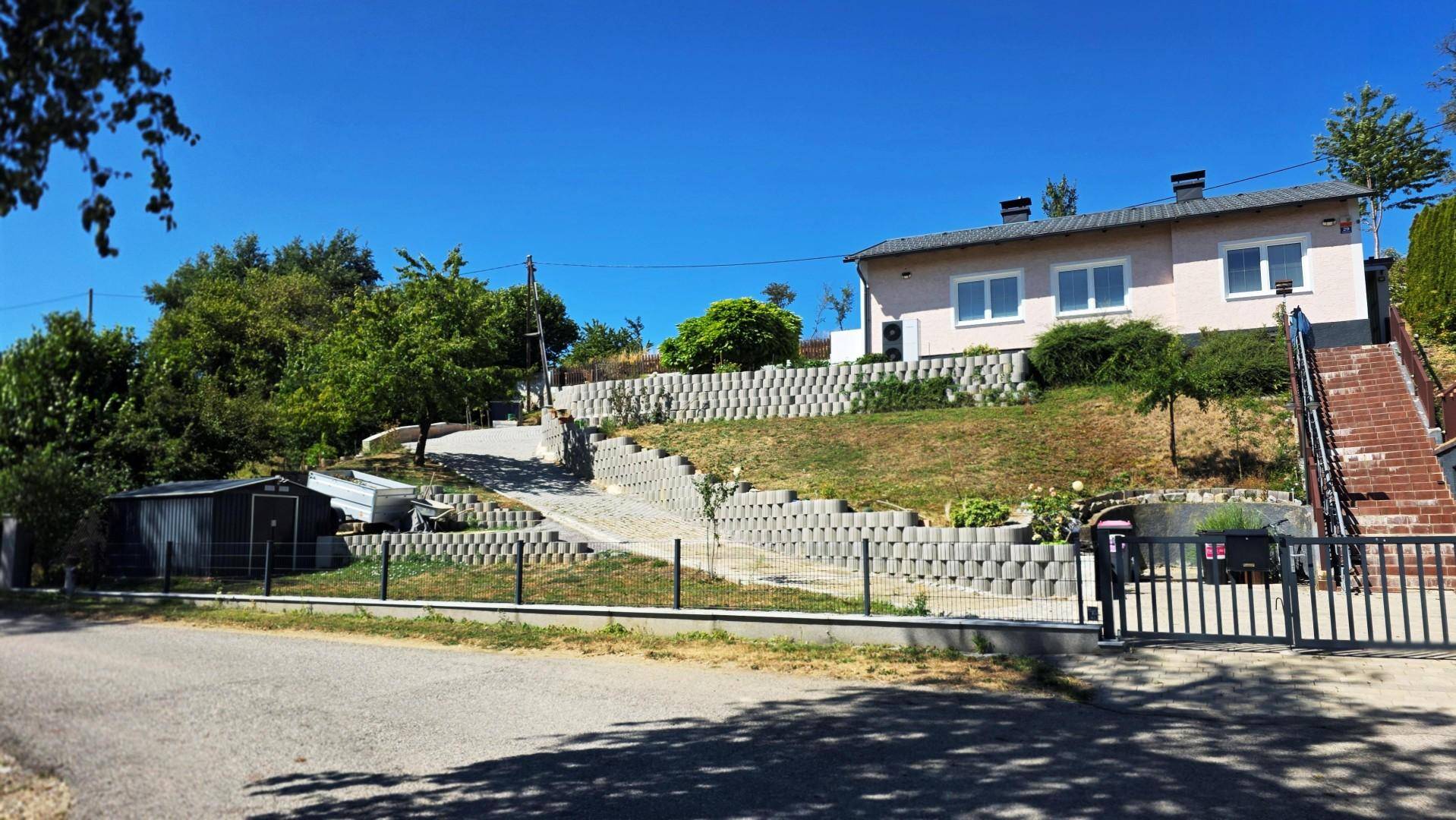 Gesamtansicht des Hauses von der Straße, mit terrassiertem Garten, Zufahrt und einem kleinen Schuppen.