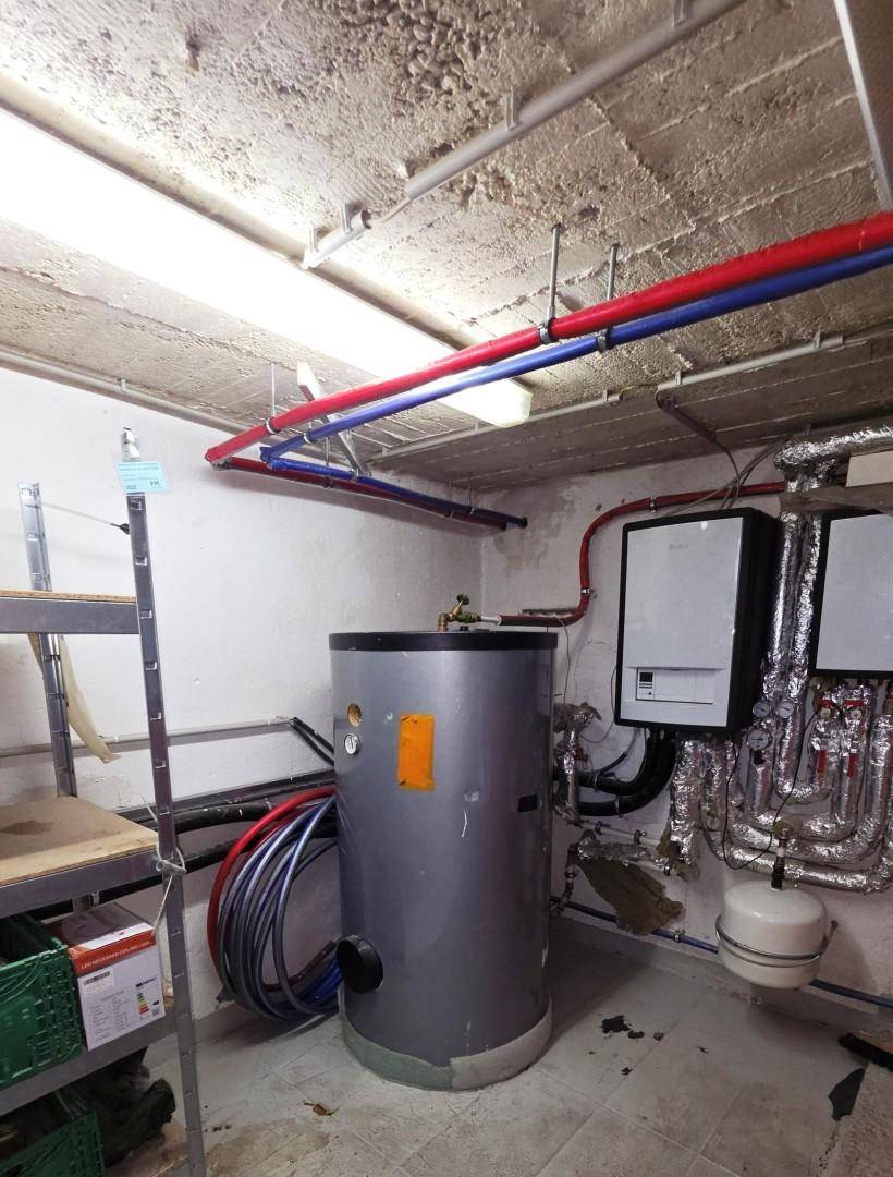 Technikraum mit Warmwasserspeicher, Rohrleitungen und Regalen für Lagerung.