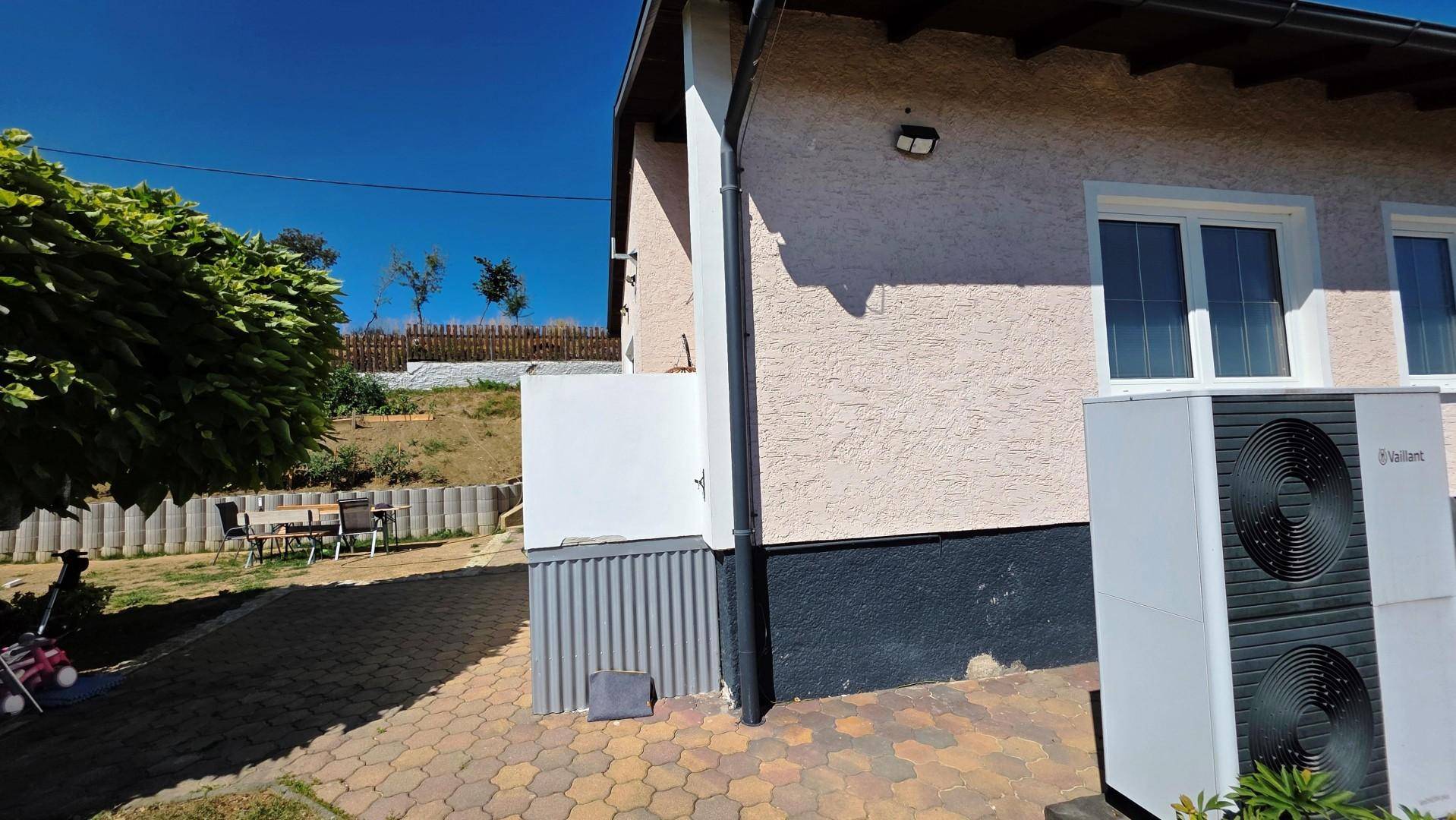 Hausaußenansicht mit Wärmepumpe, gepflastertem Weg und Blick auf eine kleine Terrasse.