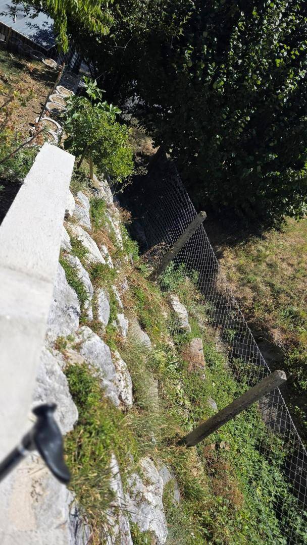 Detailansicht einer terrassierten Stützmauer mit Vegetation und einem Maschendrahtzaun im Sonnenlicht.