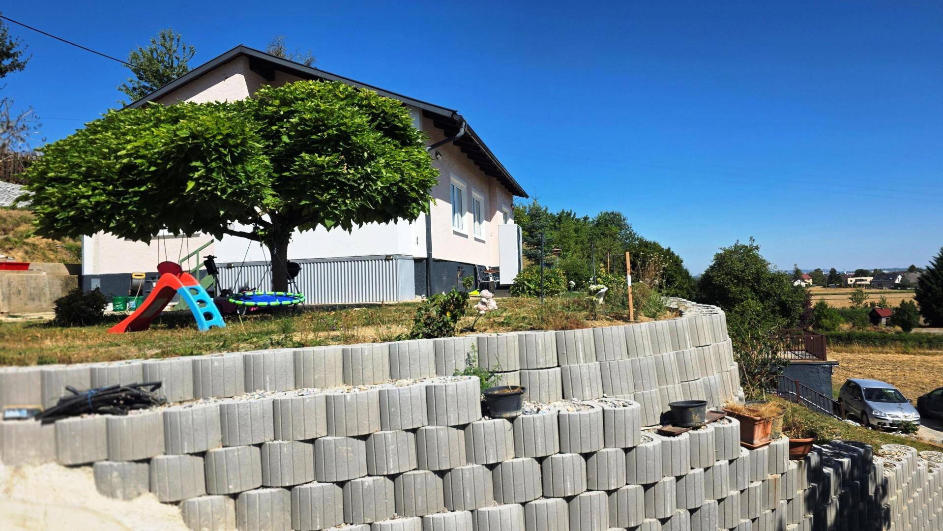Hausfassade mit terrassierter Stützmauer, Spielgeräten und weitem Blick über die Landschaft.