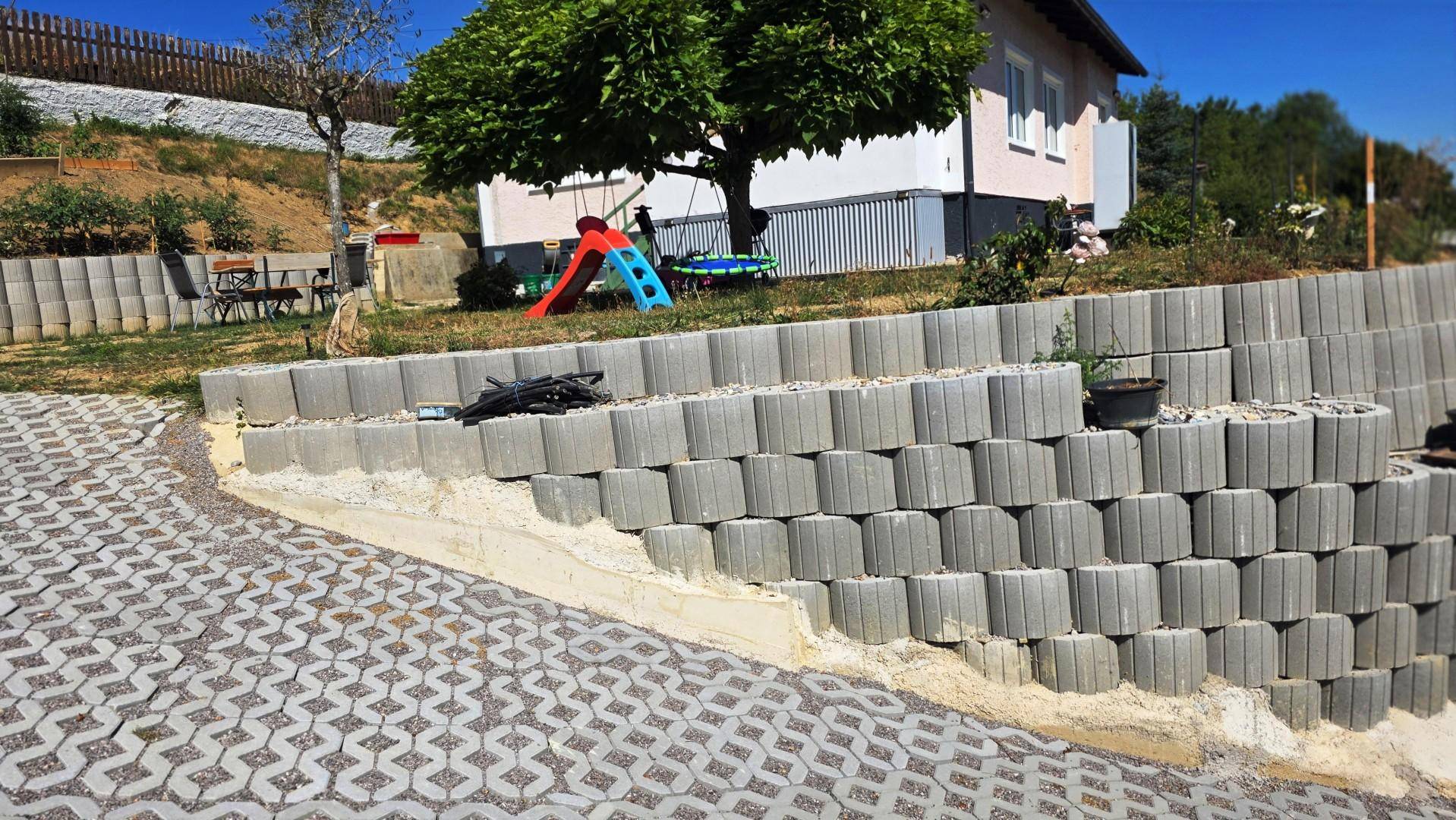 Terrassierte Stützmauer neben einem gepflasterten Bereich, mit Haus und Spielgeräten im Hintergrund.