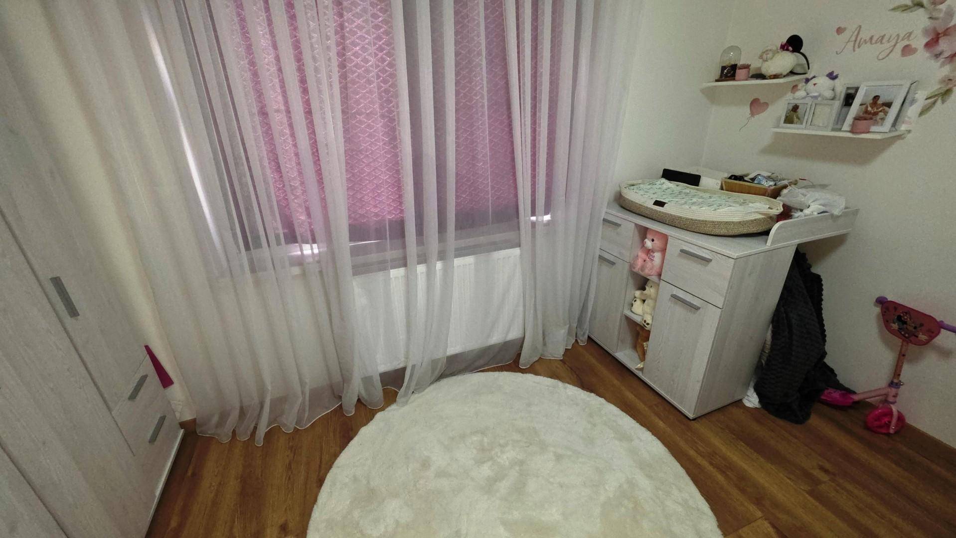 Kinderzimmer mit Wickeltisch, Holzfußboden und großem Fenster mit Vorhängen.