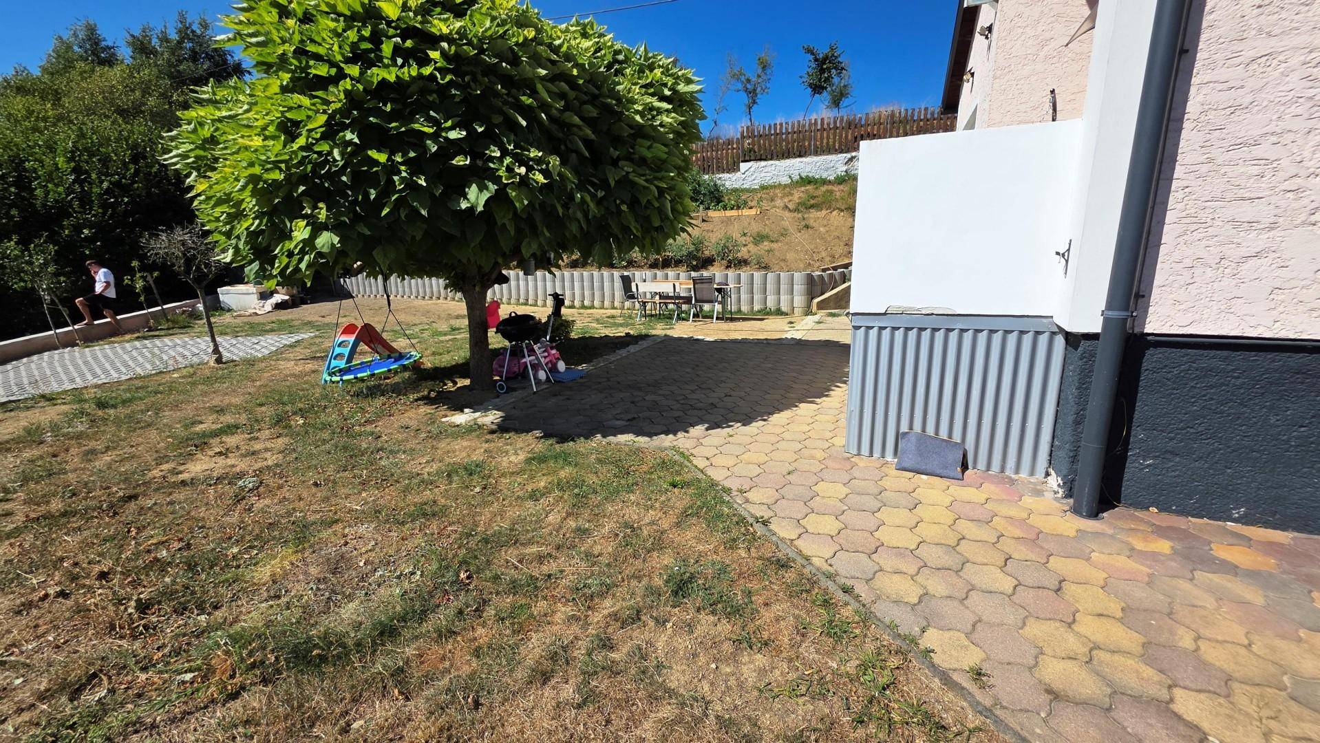 Gartenbereich mit Baum, Schaukel, Grill und gepflastertem Weg zu einer kleinen Terrasse.