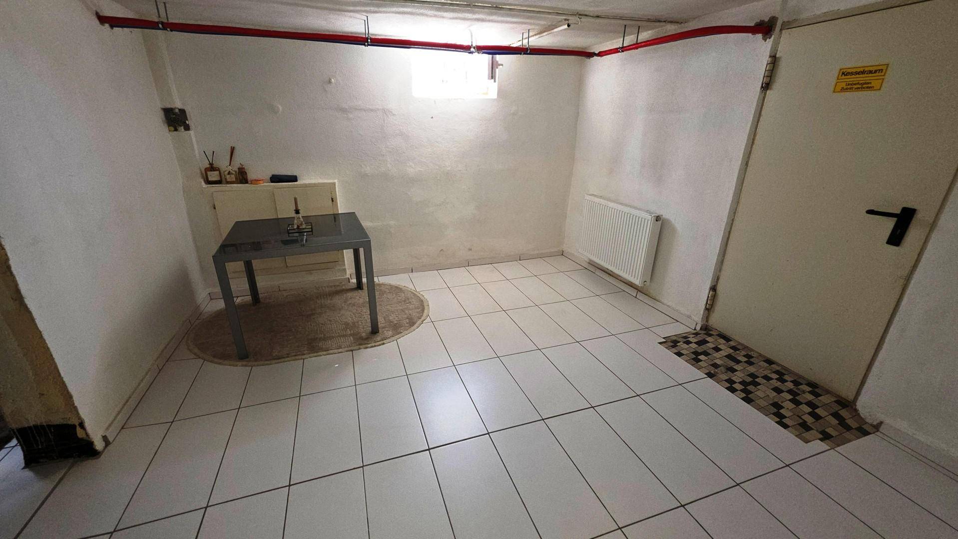 Heller Kellerraum mit weißen Fliesen, Heizkörper und sichtbaren Leitungen an der Decke.