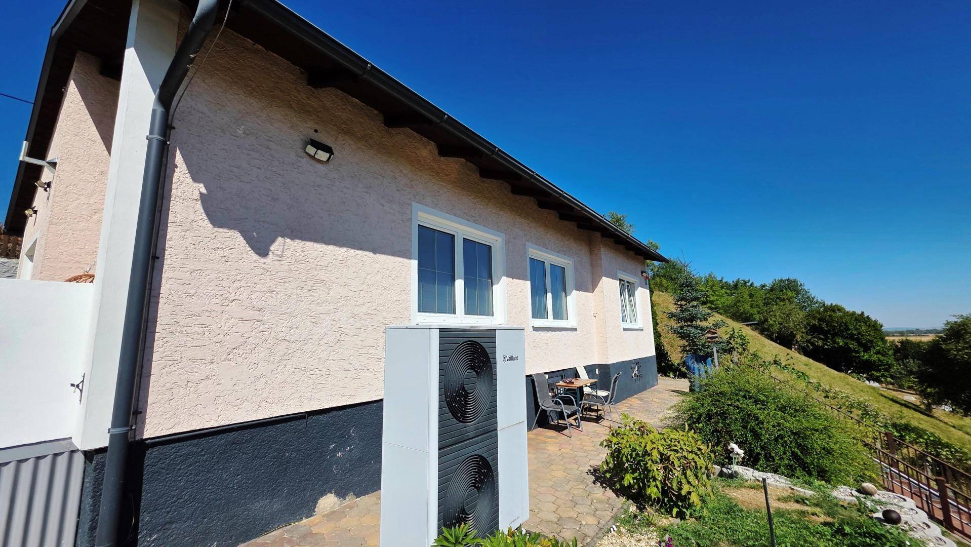 Hausfassade mit Wärmepumpe, gepflastertem Weg und kleiner Sitzgelegenheit im Grünen.