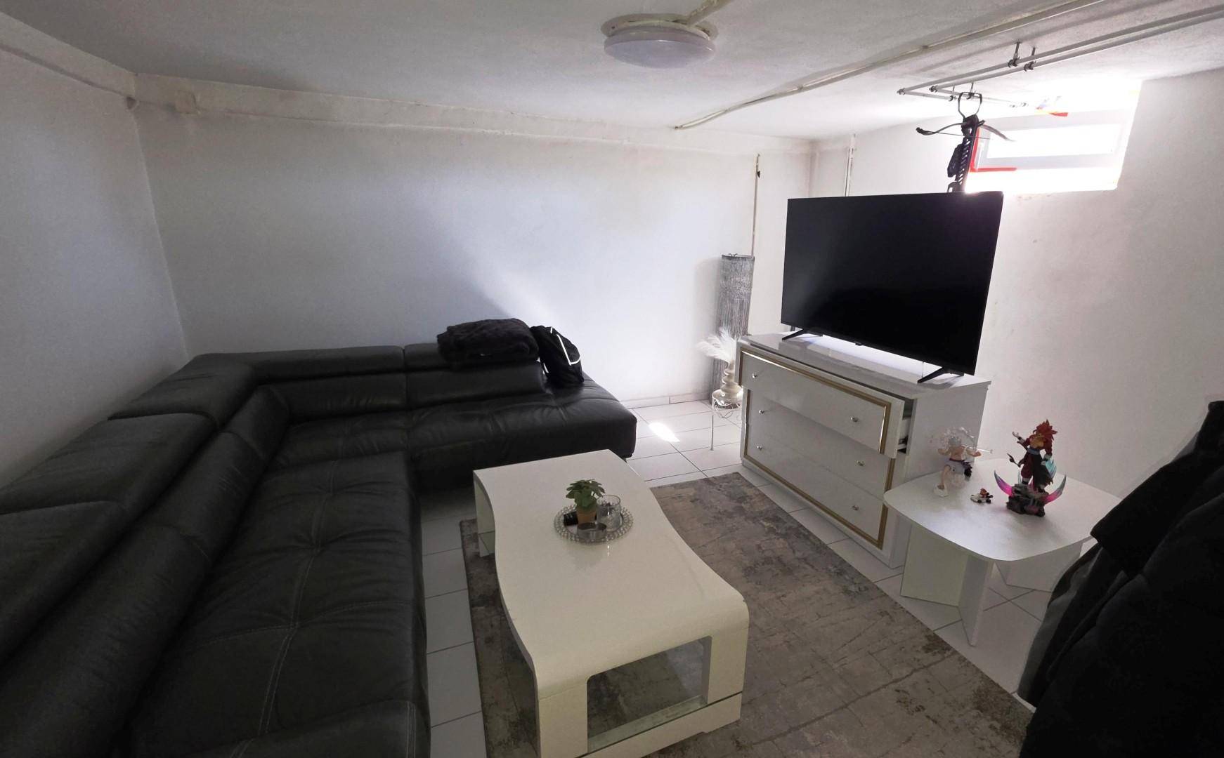Gemütlicher Wohnbereich im Keller mit schwarzem Sofa, Fernseher und weißen Fliesenboden.