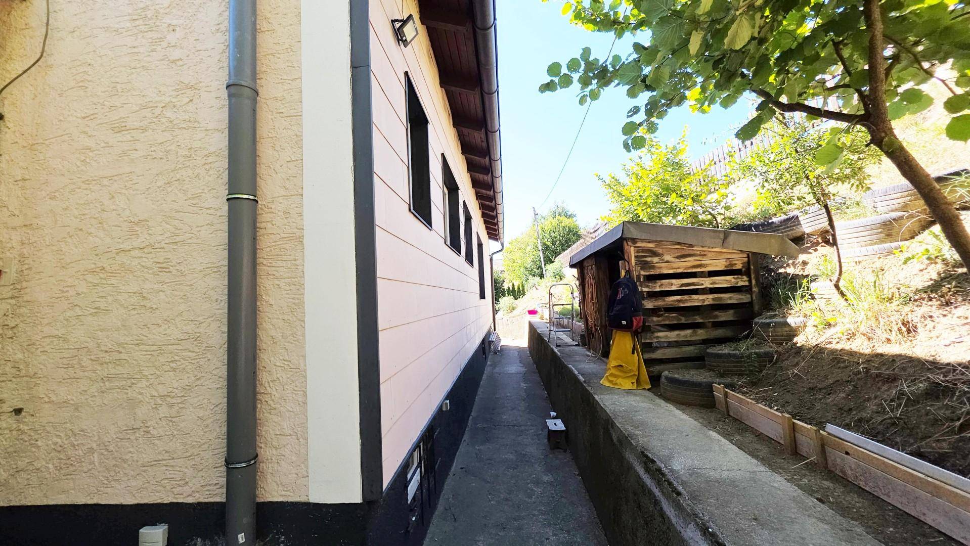 Schmaler Weg entlang der Hausseite mit kleinem Holzschuppen und Hanglage im Hintergrund.