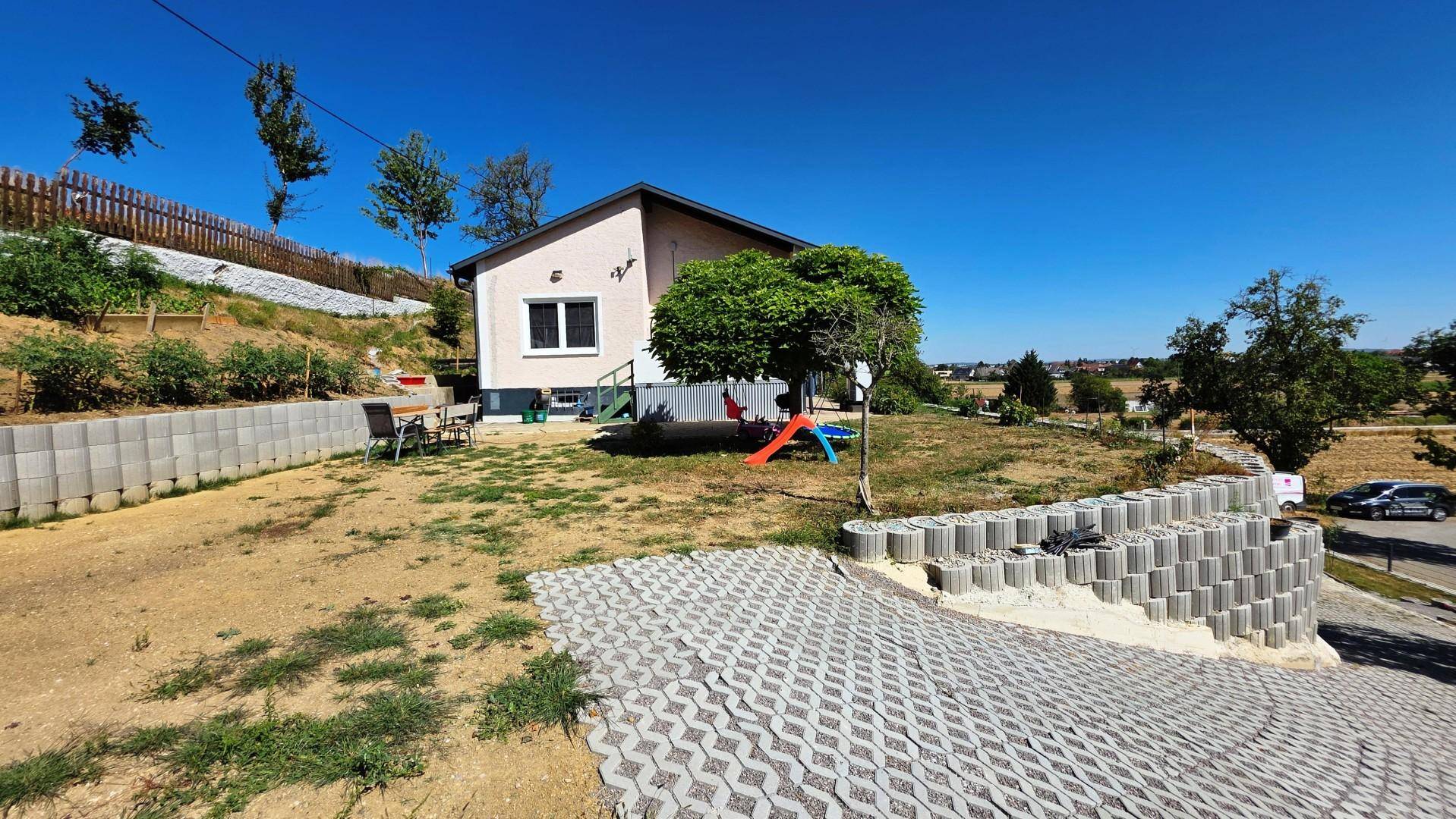 Haus mit großem Gartenbereich, Spielgeräten und terrassierten Beeten, umgeben von ländlicher Landschaft.