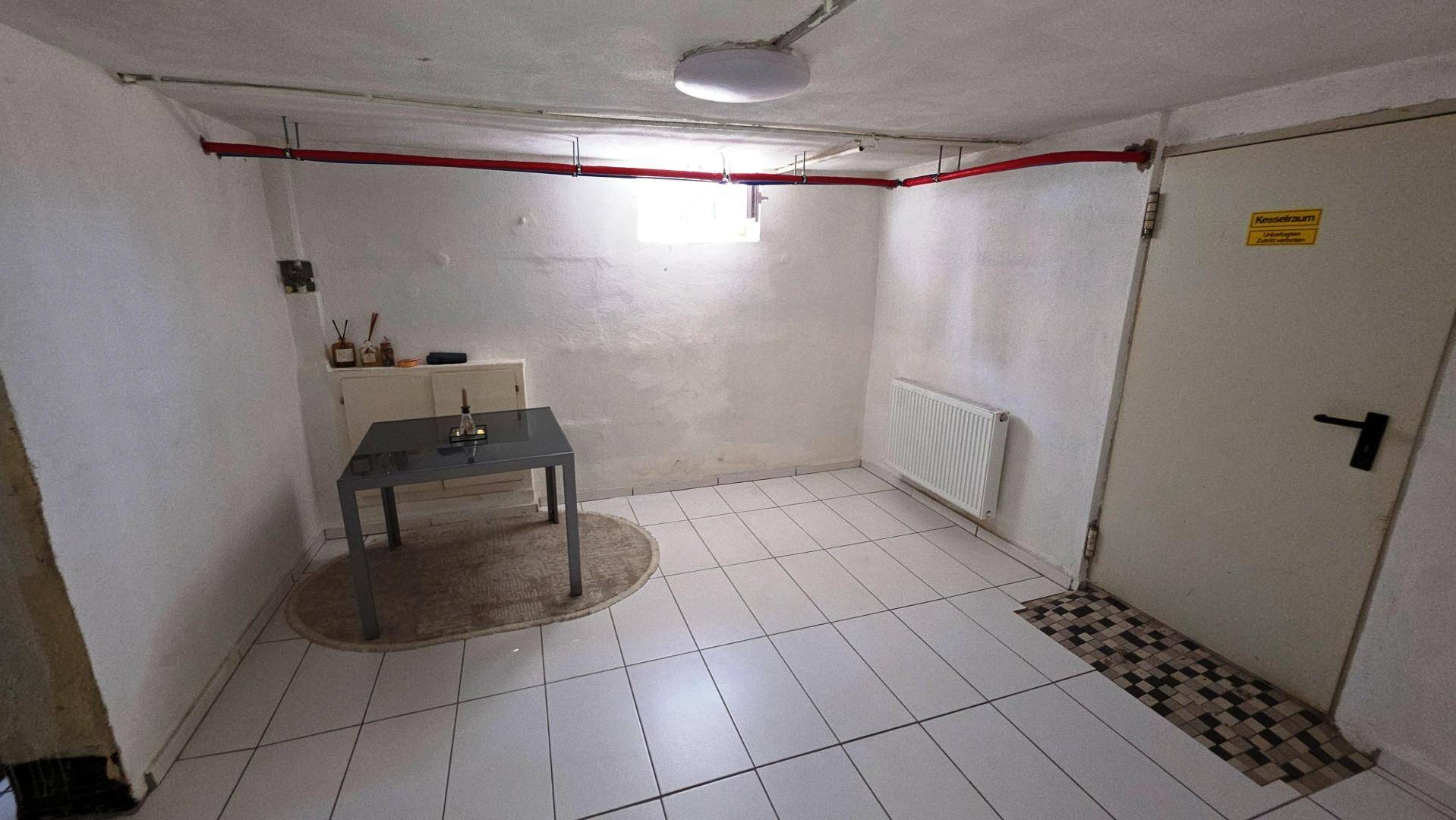 Heller Kellerraum mit weißen Fliesen, Heizkörper und sichtbaren Leitungen an der Decke.