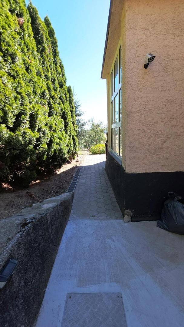 Seitenansicht des Hauses mit gepflastertem Weg, hoher Hecke und Überwachungskamera.
