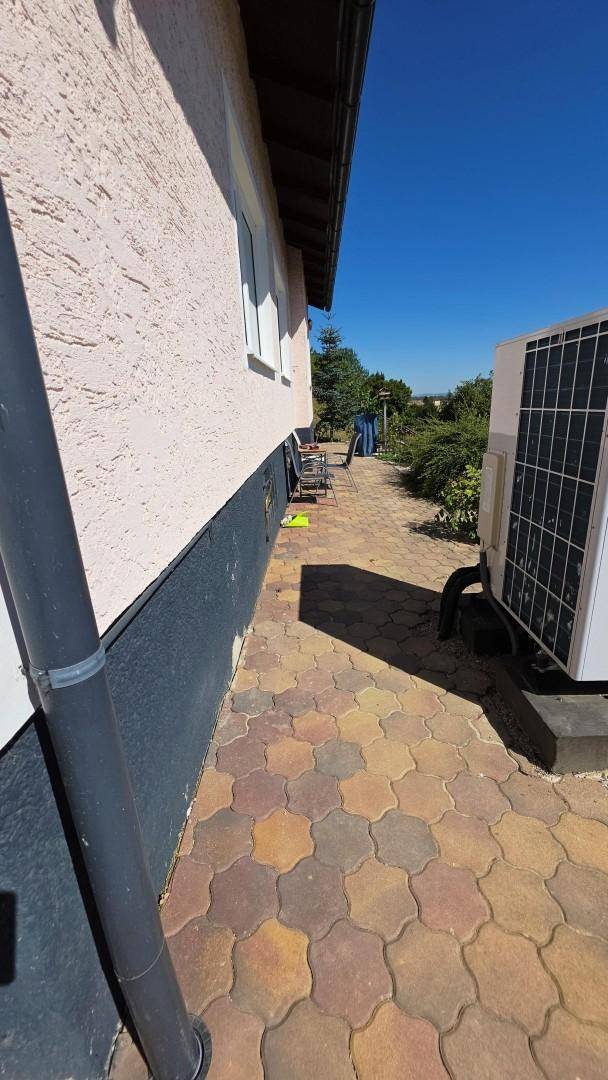 Hausseite mit gepflastertem Weg, Außeneinheit der Klimaanlage und grüner Umgebung.