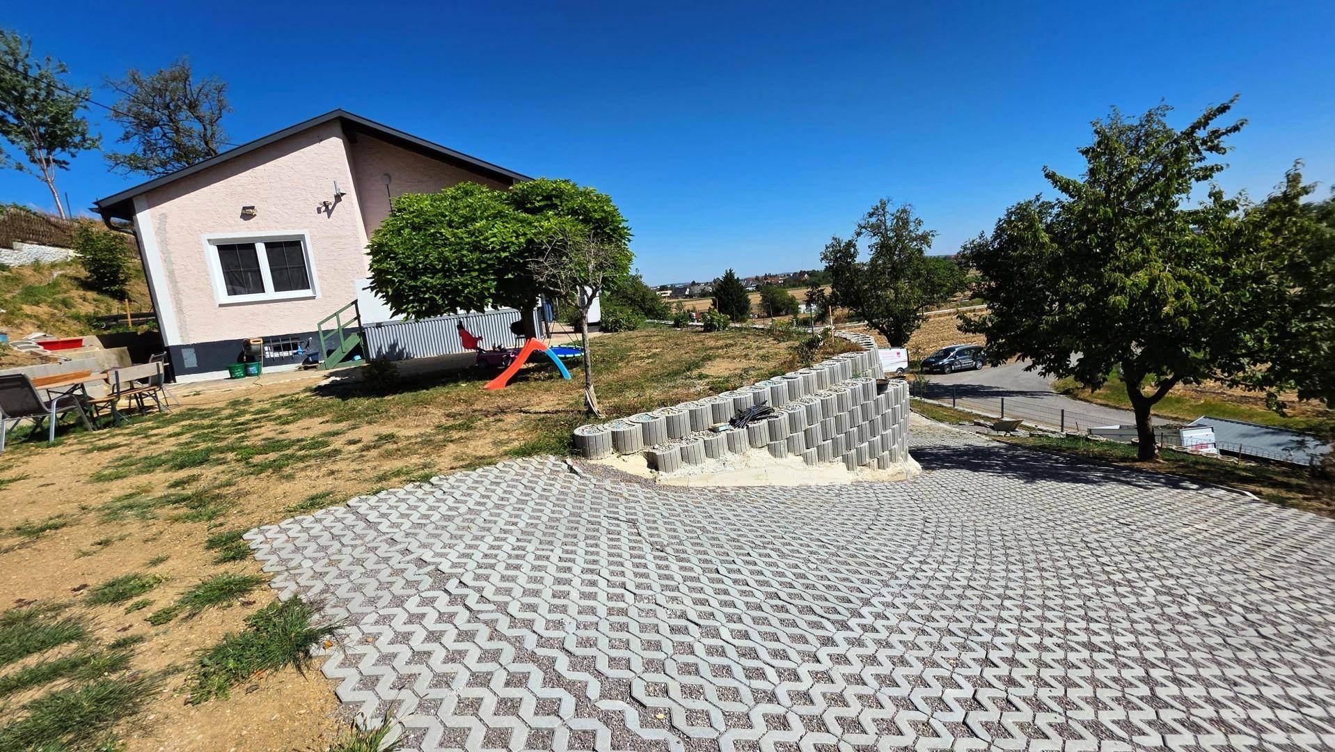 Einfamilienhaus mit gepflasterter Auffahrt, Spielbereich im Garten und terrassierten Beeten unter blauem Himmel.