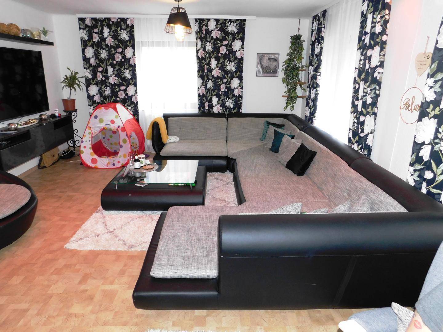 Gemütliches Wohnzimmer mit großem Ecksofa, Fernseher und floralen Vorhängen, ausgestattet mit Laminatboden.