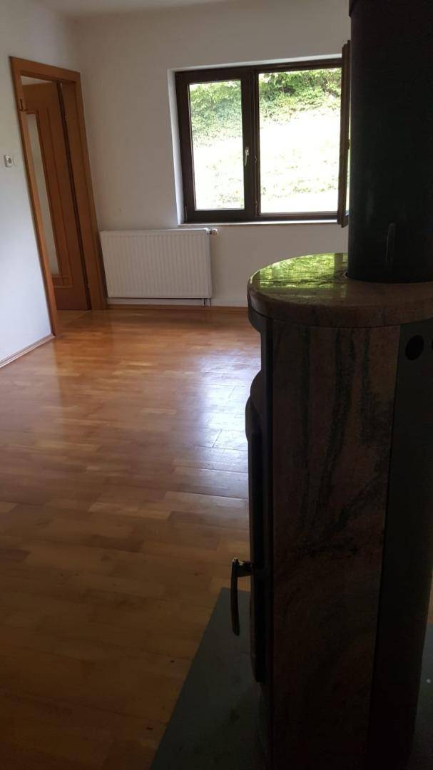 Leerer Raum mit Holzboden, Fenster und Heizkörper, ein Ofen ist teilweise sichtbar.