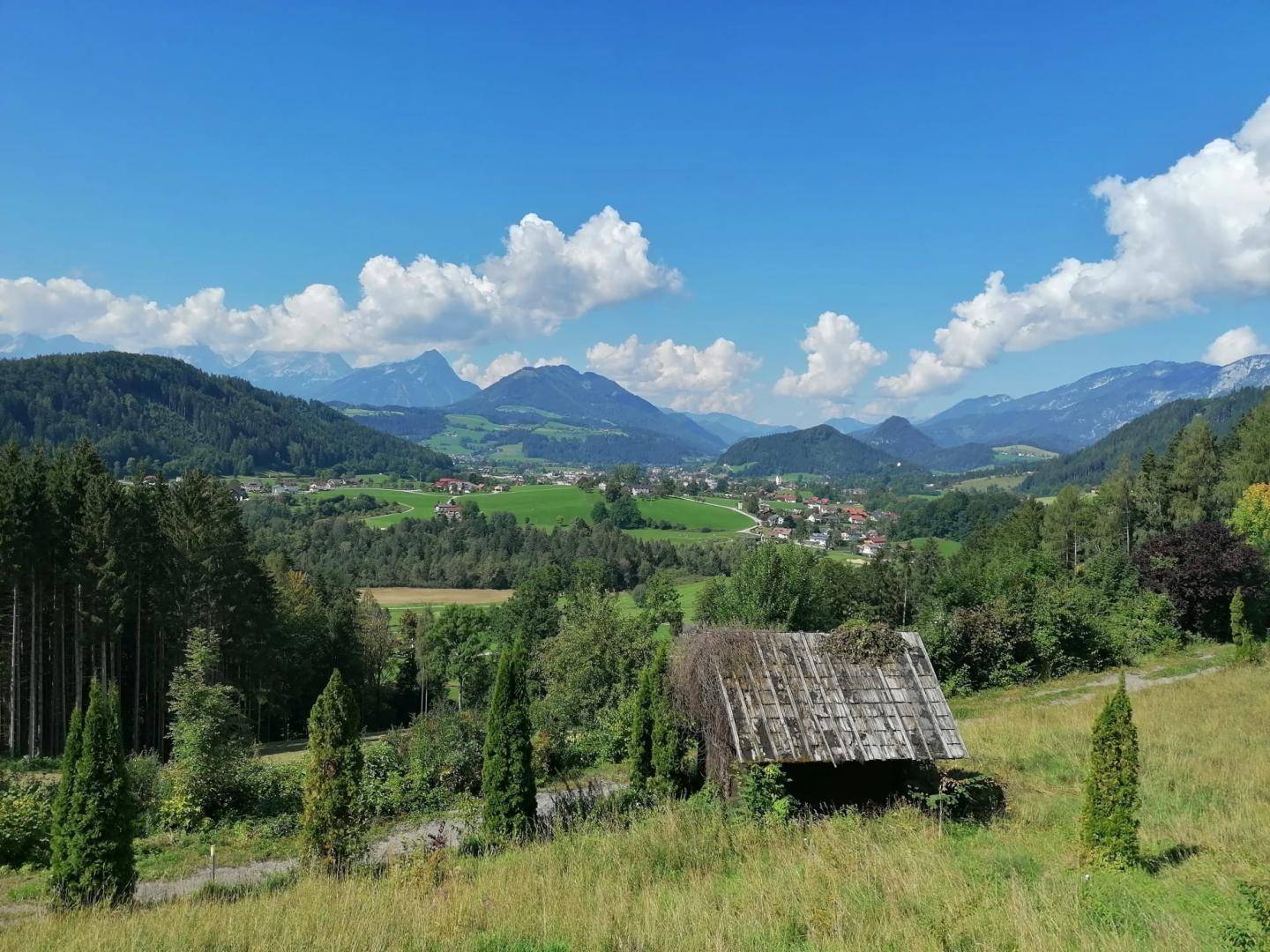 Weitläufige Landschaft mit grünen Hügeln, Bergen und einem Dorf im Tal unter blauem Himmel.