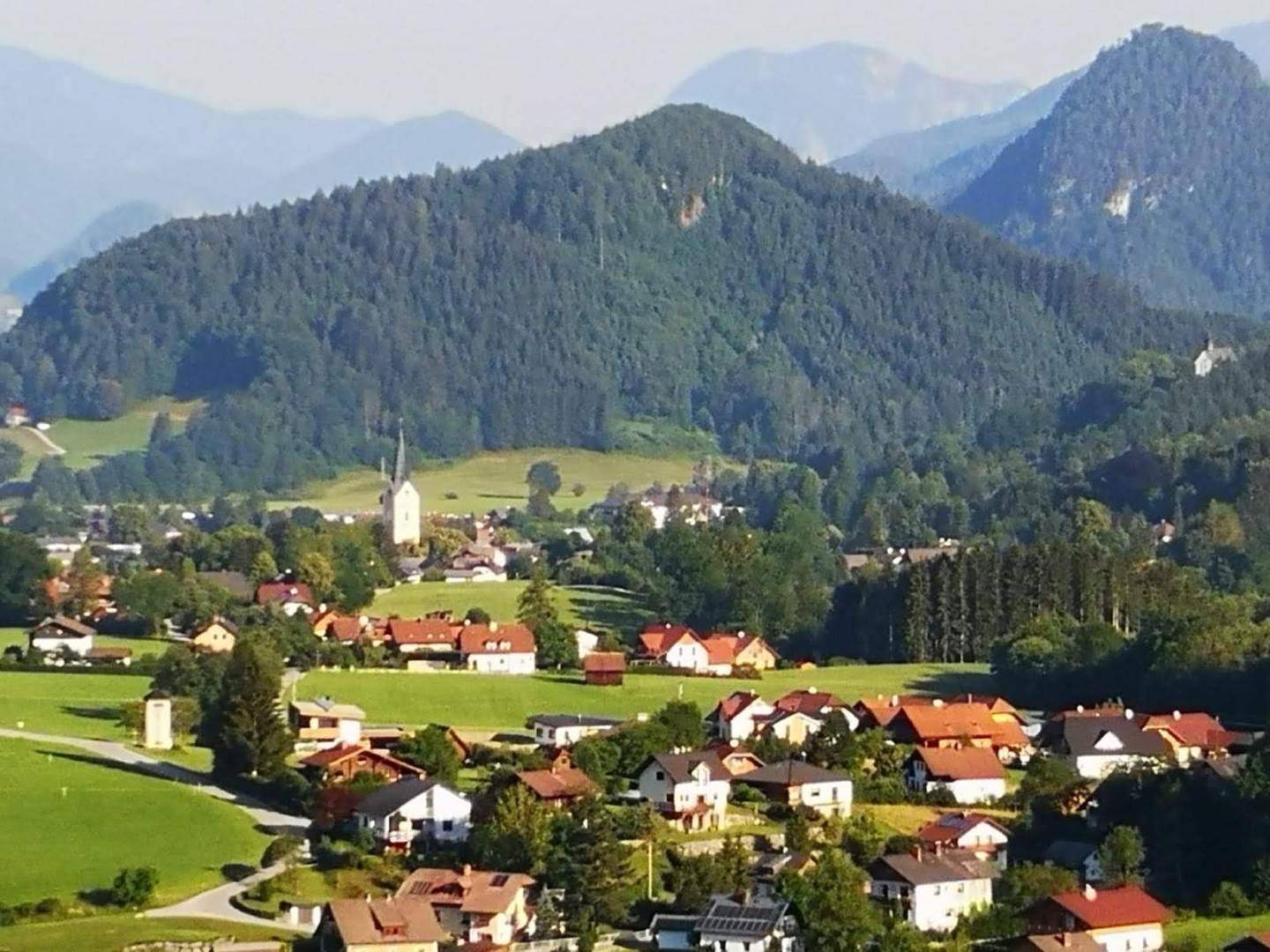 Idyllisches Dorf in einem grünen Tal, umgeben von bewaldeten Hügeln und Bergen unter blauem Himmel.