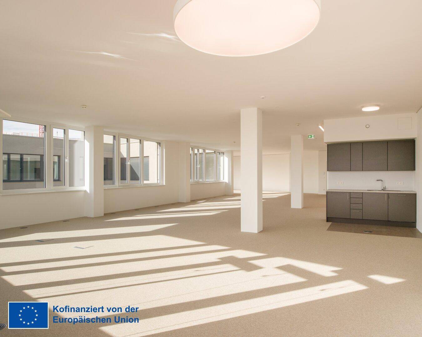 Großer, lichtdurchfluteter Büroraum mit moderner Küchenzeile, Teppichboden und weitreichenden Fenstern.