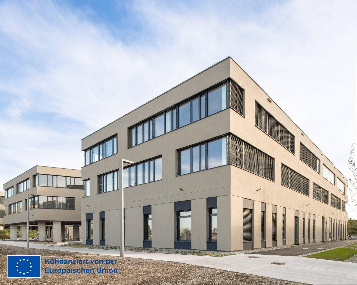 Zwei moderne Bürogebäude mit beiger Fassade, großen Fenstern und minimalistischer Landschaftsgestaltung.