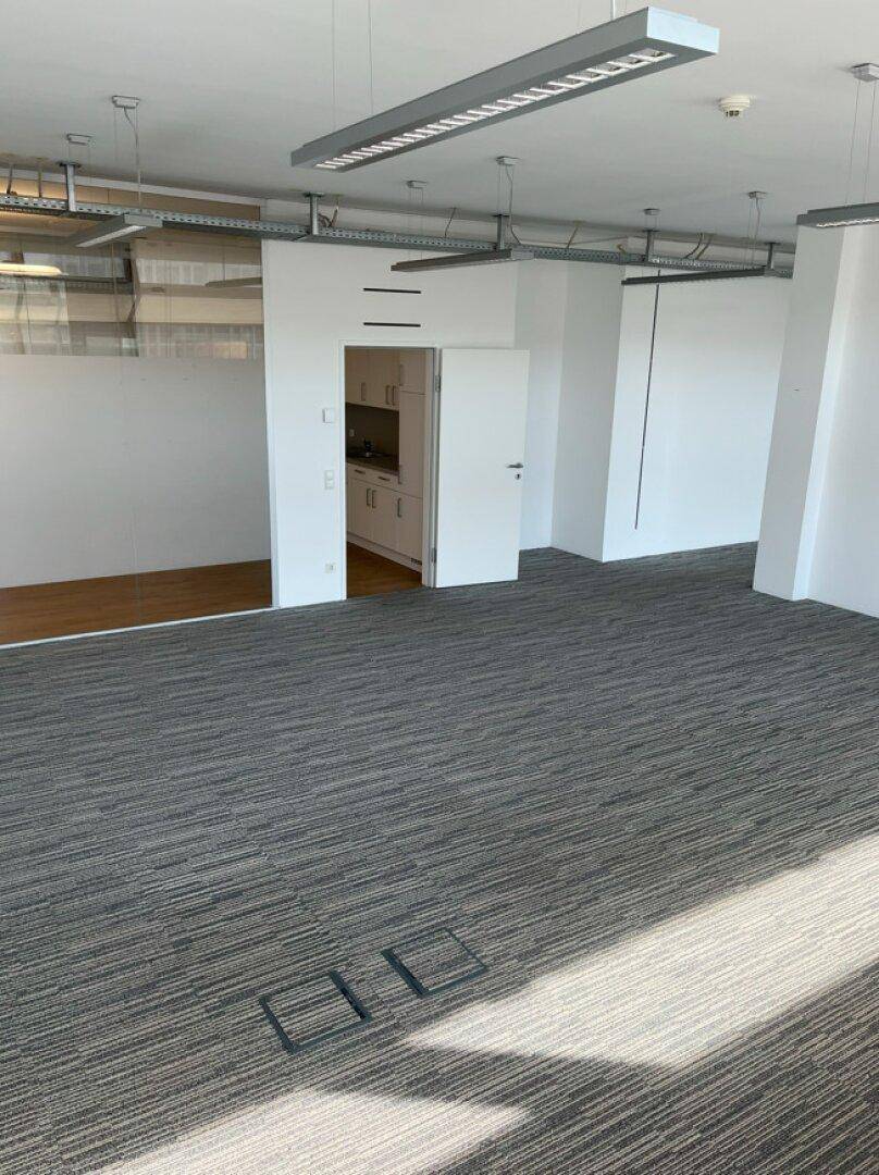 Offener Bürobereich mit Teppichboden und Zugang zu einer kleinen, funktionalen Küchenzeile.