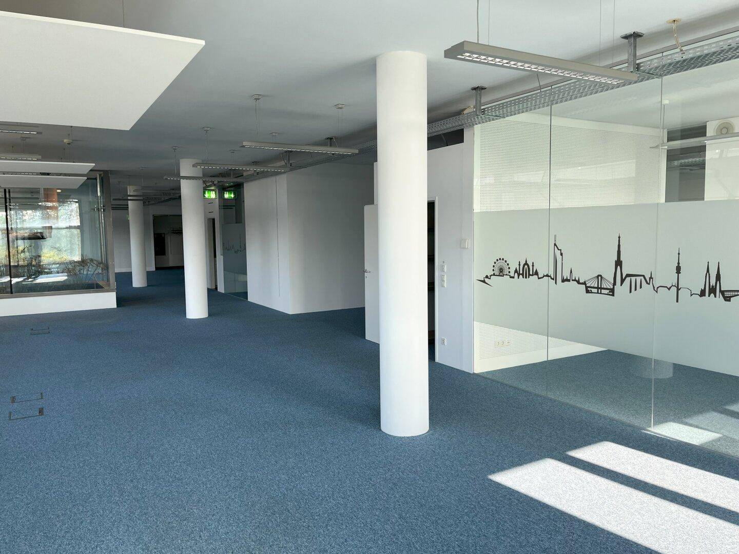 Moderner Bürobereich mit blauer Teppichboden, Säulen und Glaswänden mit Stadtmotiv.