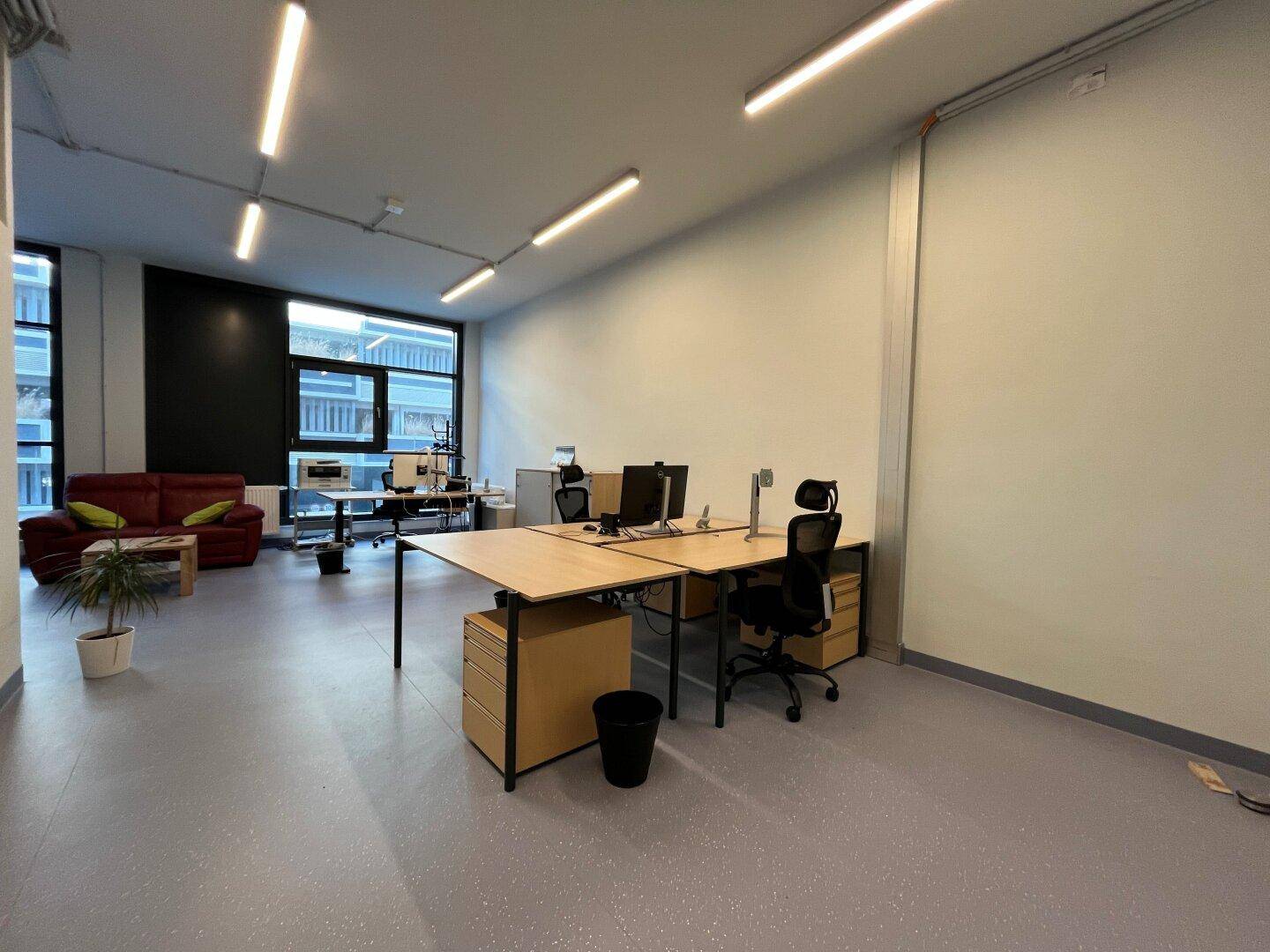 Geräumiges, modernes Büro mit mehreren Arbeitsplätzen, großen Fenstern und einem roten Sofa im Loungebereich.