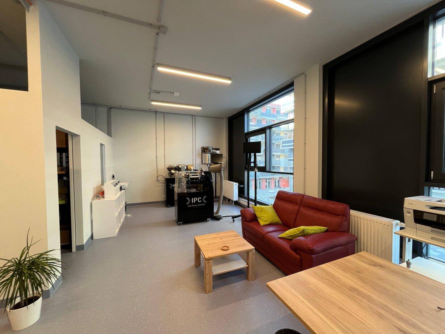 Gemütlicher Loungebereich in einem Büro mit rotem Sofa, Holztisch und großen Fenstern für viel Tageslicht.