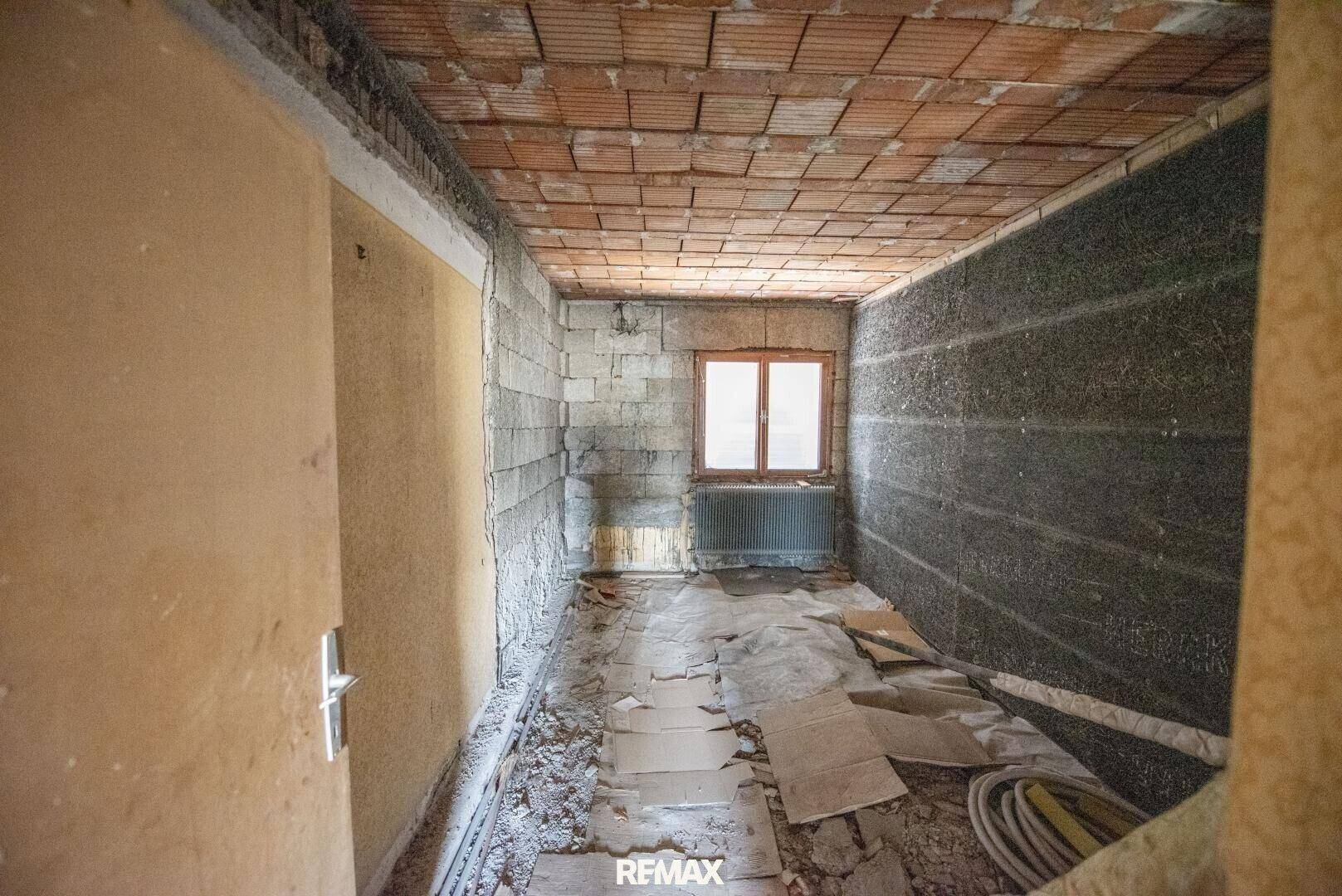 Schmaler Raum im Rohbauzustand mit Ziegelwänden und einem Fenster.