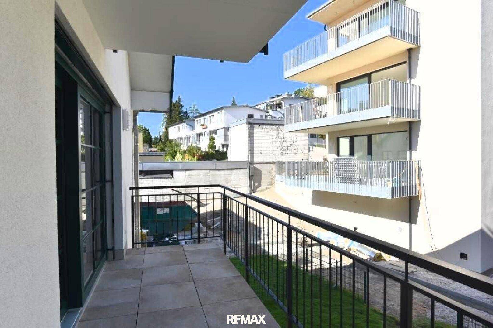 Balkon mit Fliesenboden und schwarzem Geländer, bietet Blick auf umliegende Gebäude und Grünflächen.