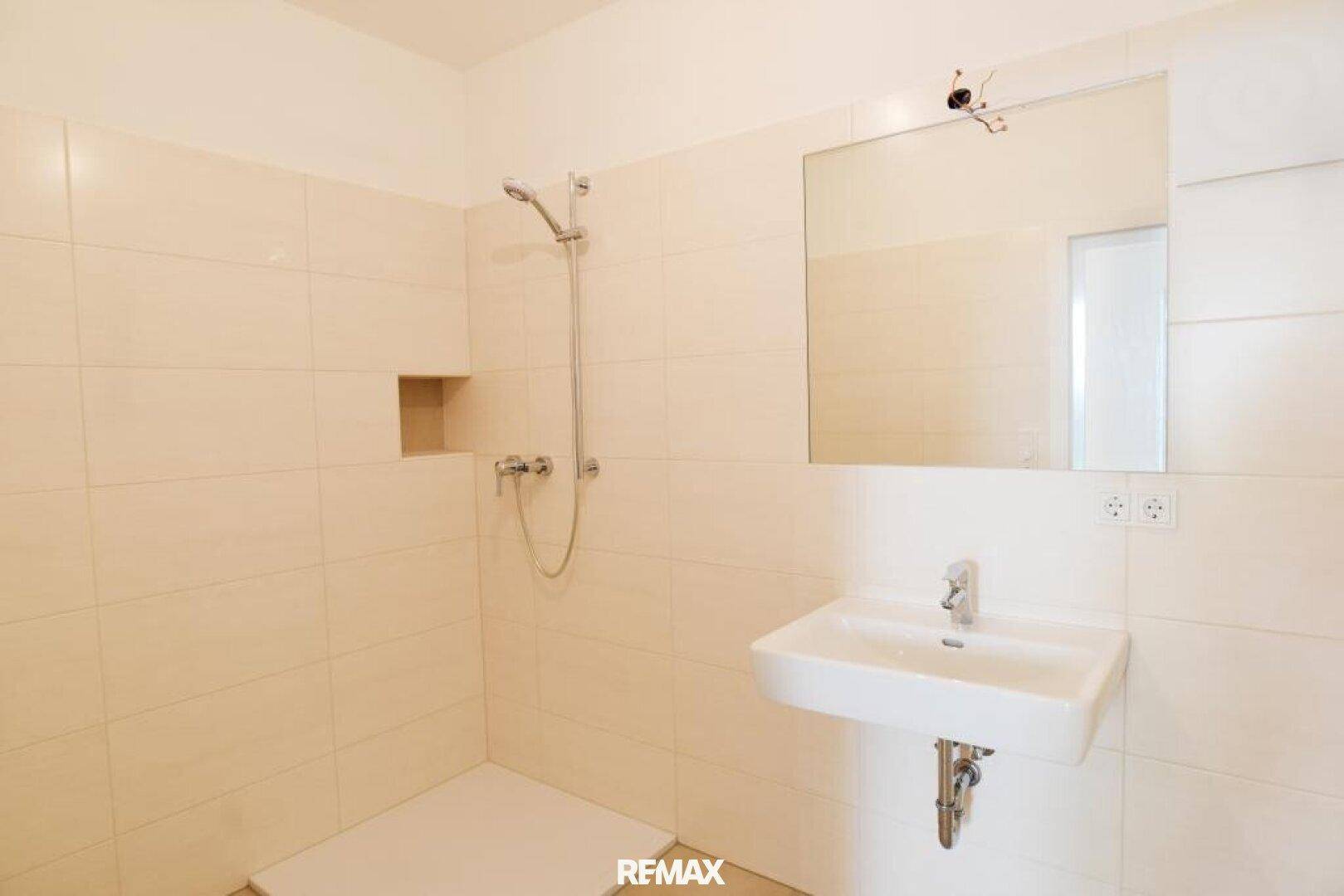 Modernes Badezimmer mit beigefarbenen Fliesen, wandmontiertem Waschbecken und Duschbereich.