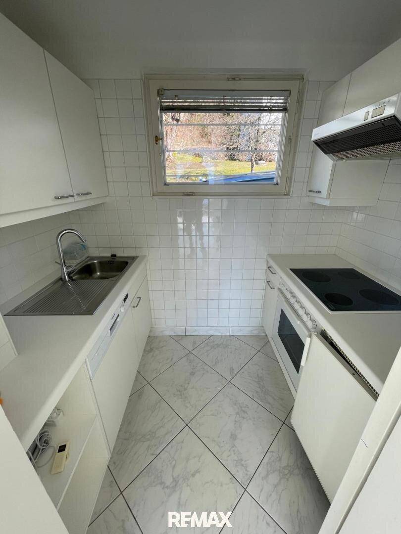 Kleine, funktionale Küche mit weißen Schränken, Fliesenboden und Fenster mit Blick ins Grüne.