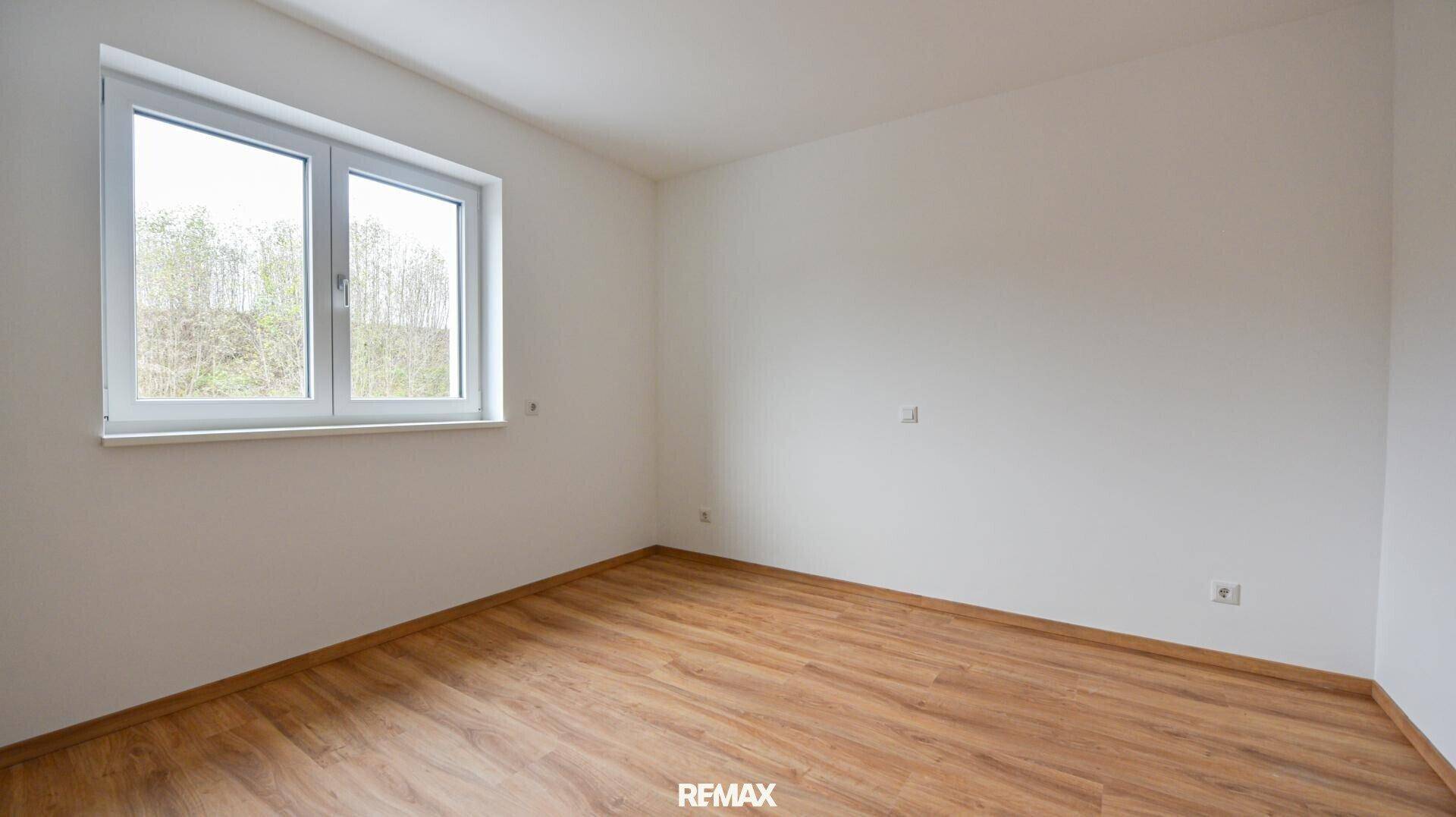 Helles, leeres Zimmer mit Laminatboden und einem Fenster, das viel Tageslicht hereinlässt.