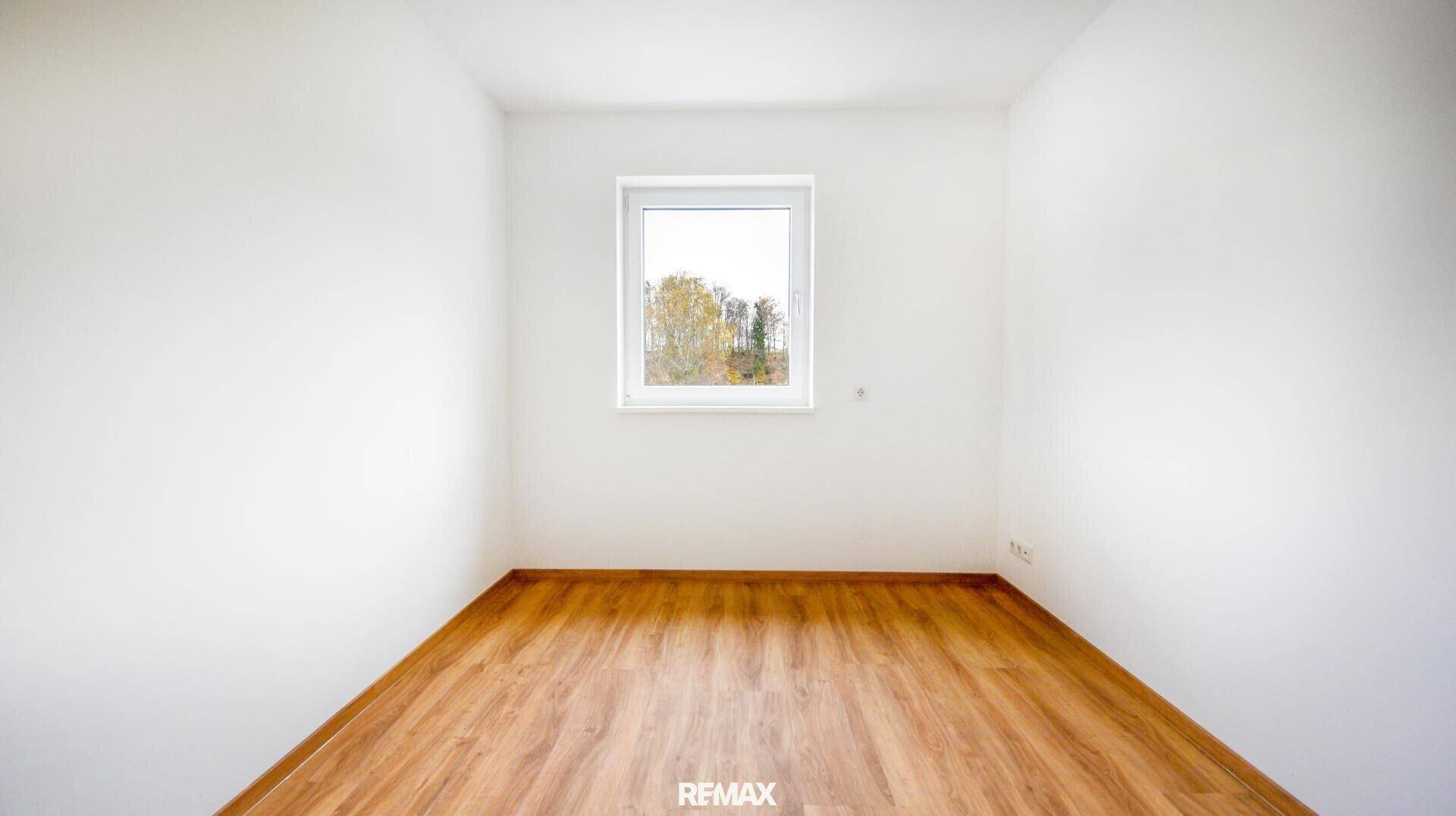 Helles, leeres Zimmer mit Laminatboden und Fenster, das einen Blick ins Grüne bietet.