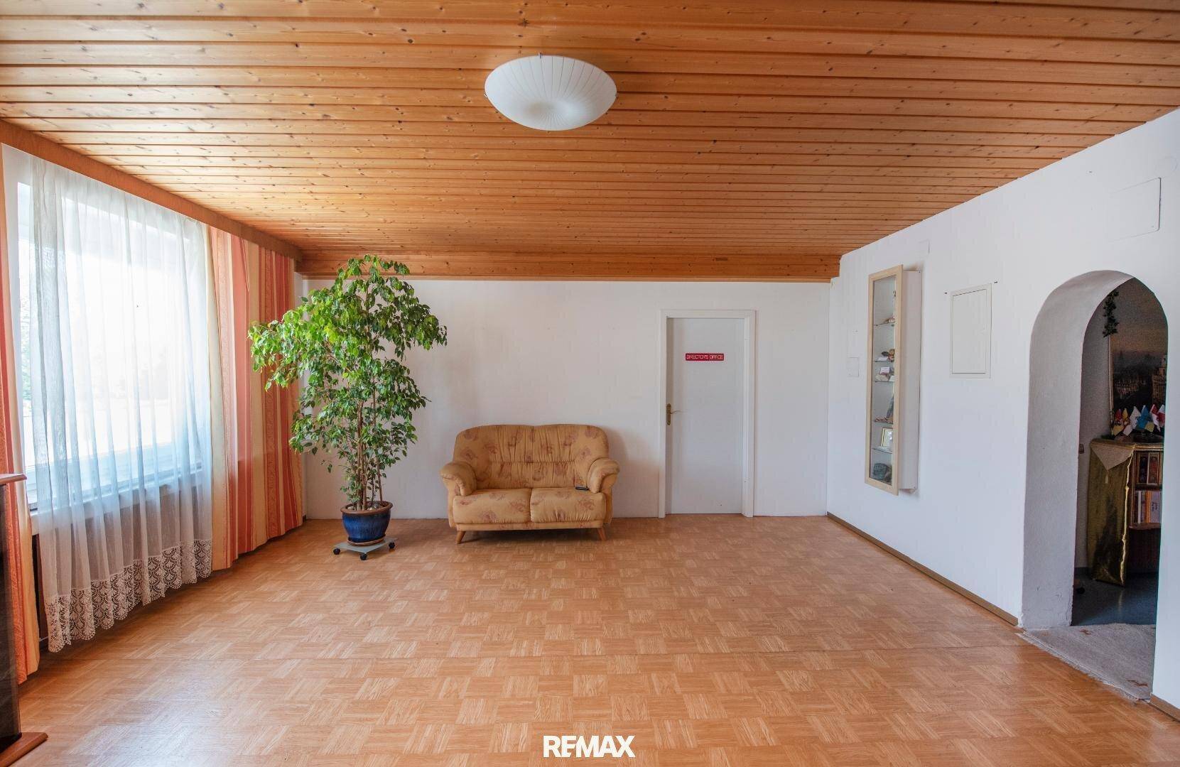 Gemütlicher Raum mit Holzdecke, gemustertem Boden, einem kleinen Sofa und einer großen Zimmerpflanze am Fenster.