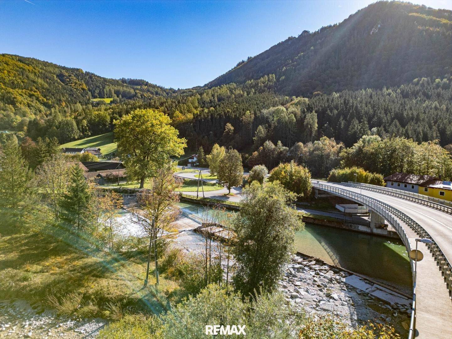 Malerische Landschaftsaufnahme mit einem Fluss, einer Brücke und bewaldeten Bergen unter blauem Himmel.