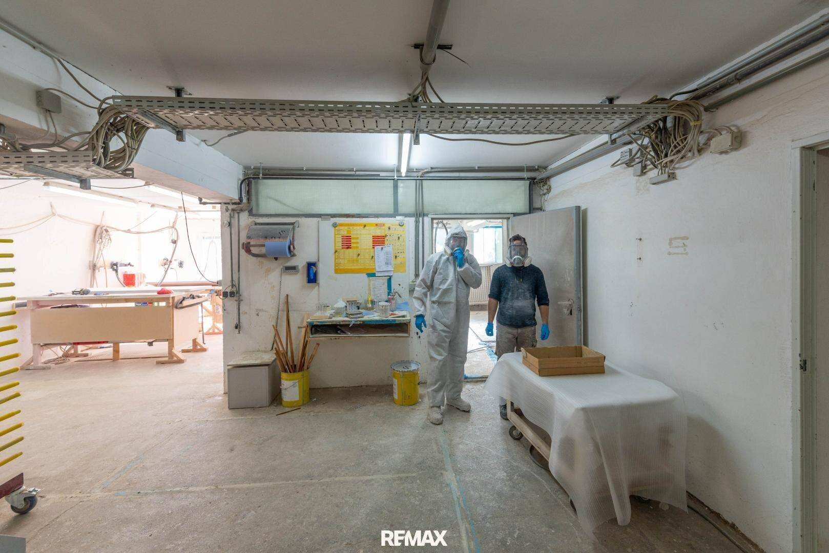 Industrieller Arbeitsbereich mit zwei Personen in Schutzkleidung und sichtbaren Kabelkanälen an der Decke.