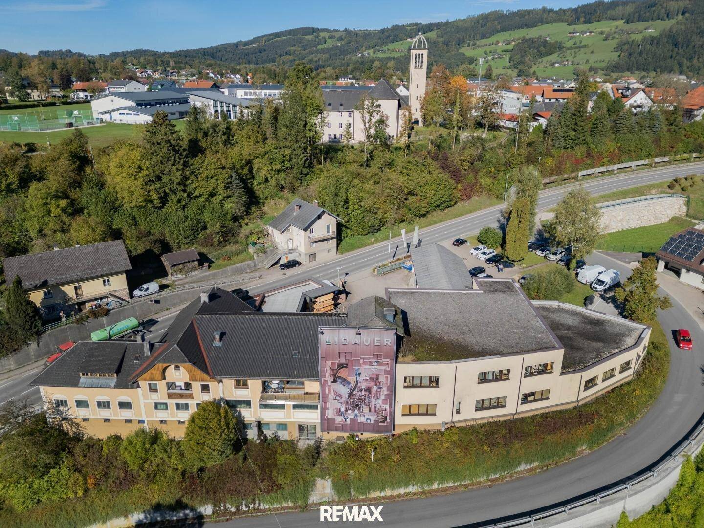 Weitläufige Luftaufnahme eines Gebäudekomplexes in einer Stadt mit Kirche und grünen Hügeln im Hintergrund.