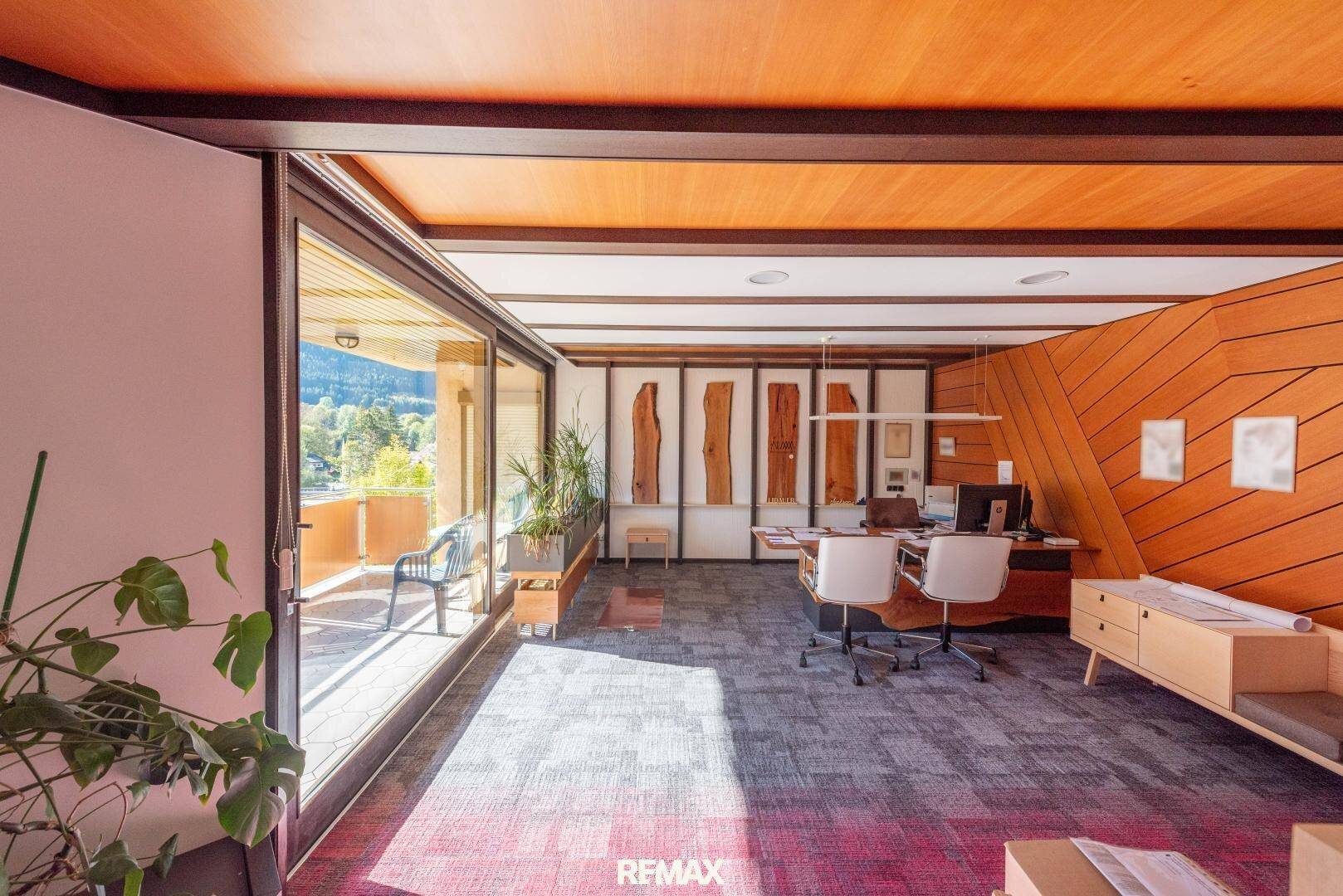 Großzügiges Büro mit Holzwänden, Holzbalkendecke, großen Fenstern und Zugang zu einem Balkon.