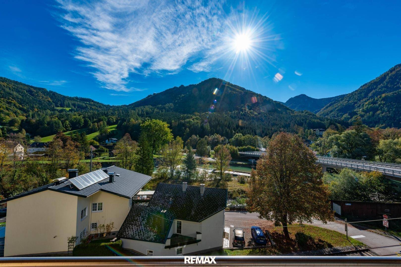Sonniger Ausblick von einem Balkon auf Häuser, eine Straße und die majestätische Berglandschaft.